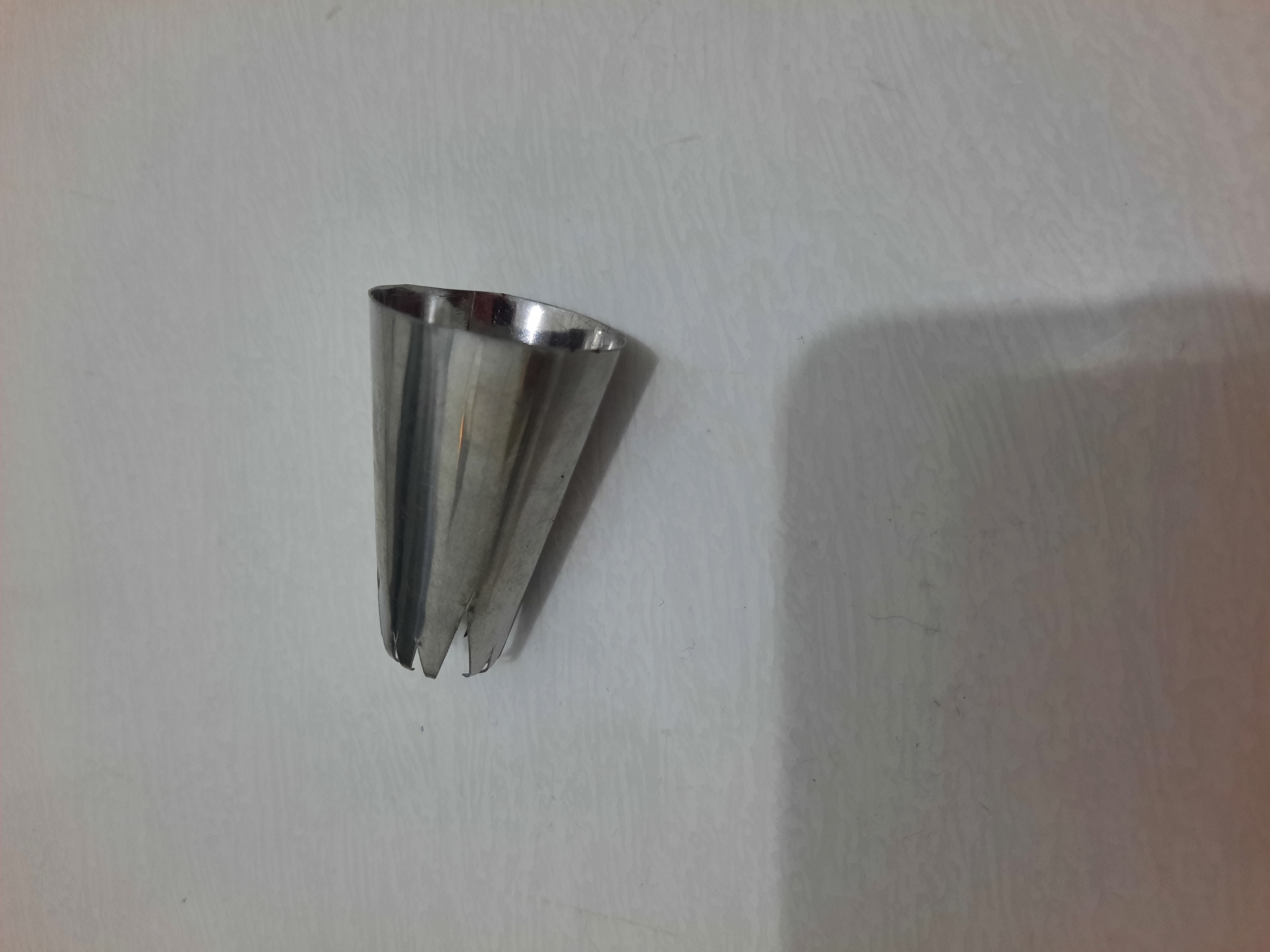 A single, metallic piping tip, shaped like a truncated cone, rests on a textured, light-colored surface. The tip features multiple slits at its wider end. The surface appears to be a table or countertop, possibly in a kitchen environment. The lighting casts a soft shadow to the left of the piping tip, indicating a light source from the right. There are no people or other objects visible in the image. The location is not identifiable from the visual cues provided.