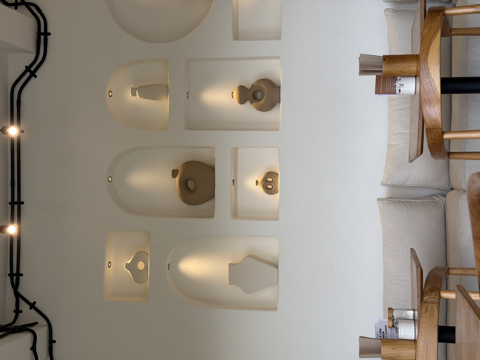 The image depicts an indoor scene, likely a cafe or restaurant in Mengwi, Indonesia, characterized by a minimalist aesthetic. The focal point is a white wall adorned with several recessed shelves, each illuminated by a spotlight from above. These shelves house decorative ceramic vases of various shapes and muted earthy tones. Above the shelves, black electrical conduits are routed across the ceiling, with two small spotlight fixtures visible. In the foreground, two wooden tables are partially visible, set with what appear to be condiments and napkins in wooden holders, suggesting a dining area. Beige upholstered seating is visible behind the tables. The overall ambiance is serene and contemporary.