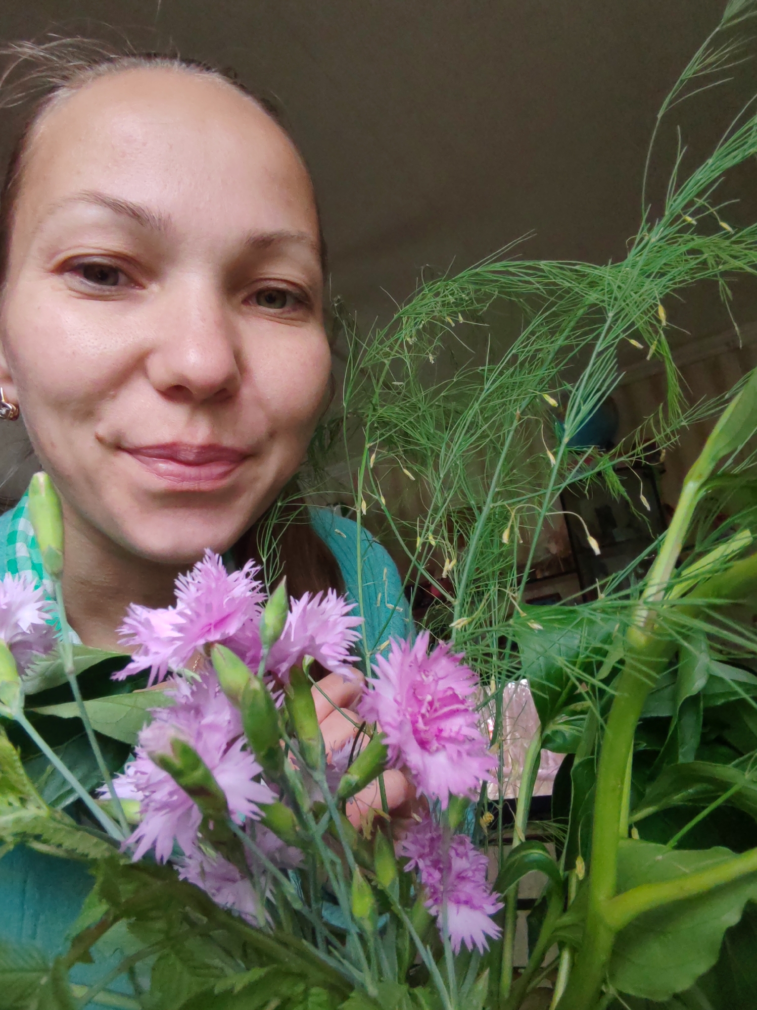 A medium shot captures a woman holding a bouquet of flowers. She is in the foreground, smiling slightly, with a green and white checkered shirt partially visible. The flowers, predominantly pink carnations and feathery green foliage, are in front of her. The background is a dark, indistinct interior. The woman's skin appears fair, with light brown eyes and light brown hair pulled back. A silver earring is visible in her left ear. The lighting suggests an indoor setting, possibly near a window given the soft light on her face. The scene appears to be within a residential area of Chuchlam, Russia.