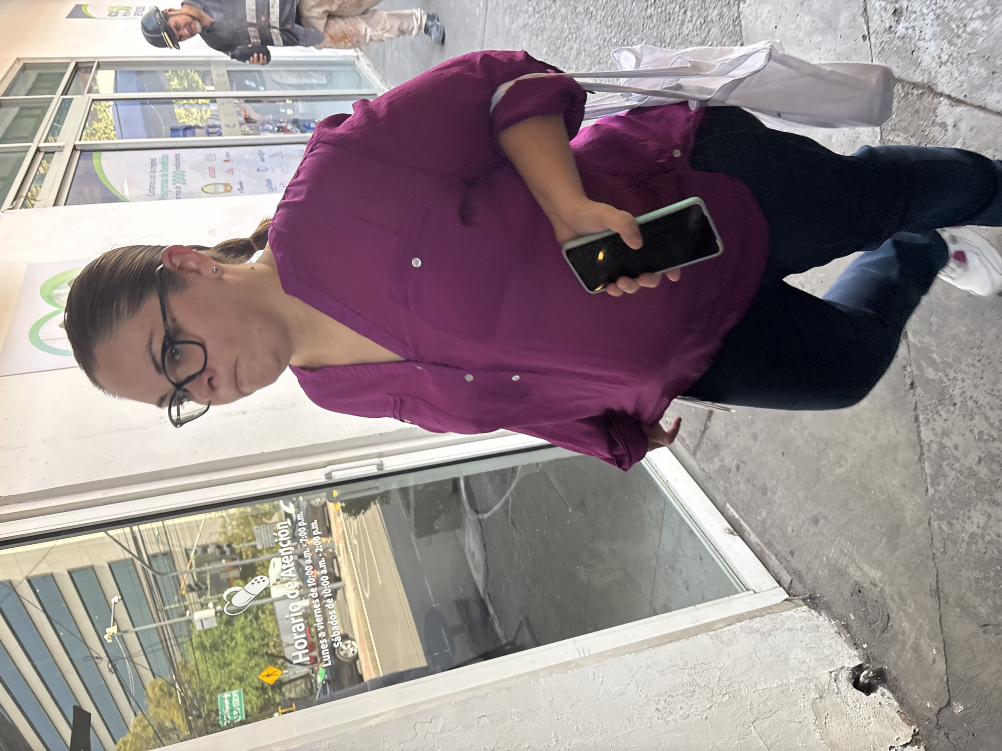 A woman stands on a concrete sidewalk in what appears to be Mexico City, Mexico, positioned in front of a building with large glass windows. She is wearing a purple long-sleeved button-up shirt, dark blue pants, and white sneakers with a pink detail. Her dark hair is pulled back, and she wears black-rimmed glasses, looking towards the camera with a neutral expression. In her right hand, she holds a black smartphone with a light-colored case. A white bag is visible, hanging from her left shoulder.
In the background, further down the sidewalk, a man in a work uniform, including a dark hard hat and possibly a reflective vest over a dark shirt, is visible walking away from the viewer. The glass windows of the building reflect trees, streetlights, and other urban structures, suggesting a bustling street scene opposite.
The image captures a daytime scene under clear weather conditions. Prominently displayed on the glass window, in Spanish, are the "Horario de Atención" (Hours of Operation): "Lunes a viernes de 10:00 a.m. - 7:00 p.m." (Monday to Friday from 10:00 a.m. - 7:00 p.m.) and "Sábados de 10:00 a.m. - 2:00 p.m." (Saturdays from 10:00 a.m. - 2:00 p.m.). Partially visible text also reads "Contamos con los mejores programas de" (We have the best programs of) and "desde 1,000" (from 1,000). The setting is an typical urban commercial area.