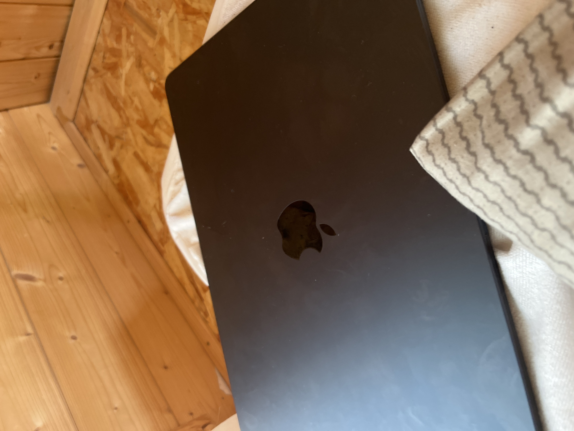 The image shows a closed, black MacBook laptop with a reflective Apple logo on its lid. The laptop is resting on a white, textured surface, possibly a blanket or cushion, which is in turn placed on a wooden floor. The background consists of angled wooden planks, suggesting an attic or a room with a sloped ceiling. The lighting in the image is warm and appears to come from above and to the right, casting subtle reflections on the laptop's surface. There are no people visible in the image. The scene is static and indoors. The location in the city of Vasilyevo, Russia, is not discernible from the visual elements of the image.