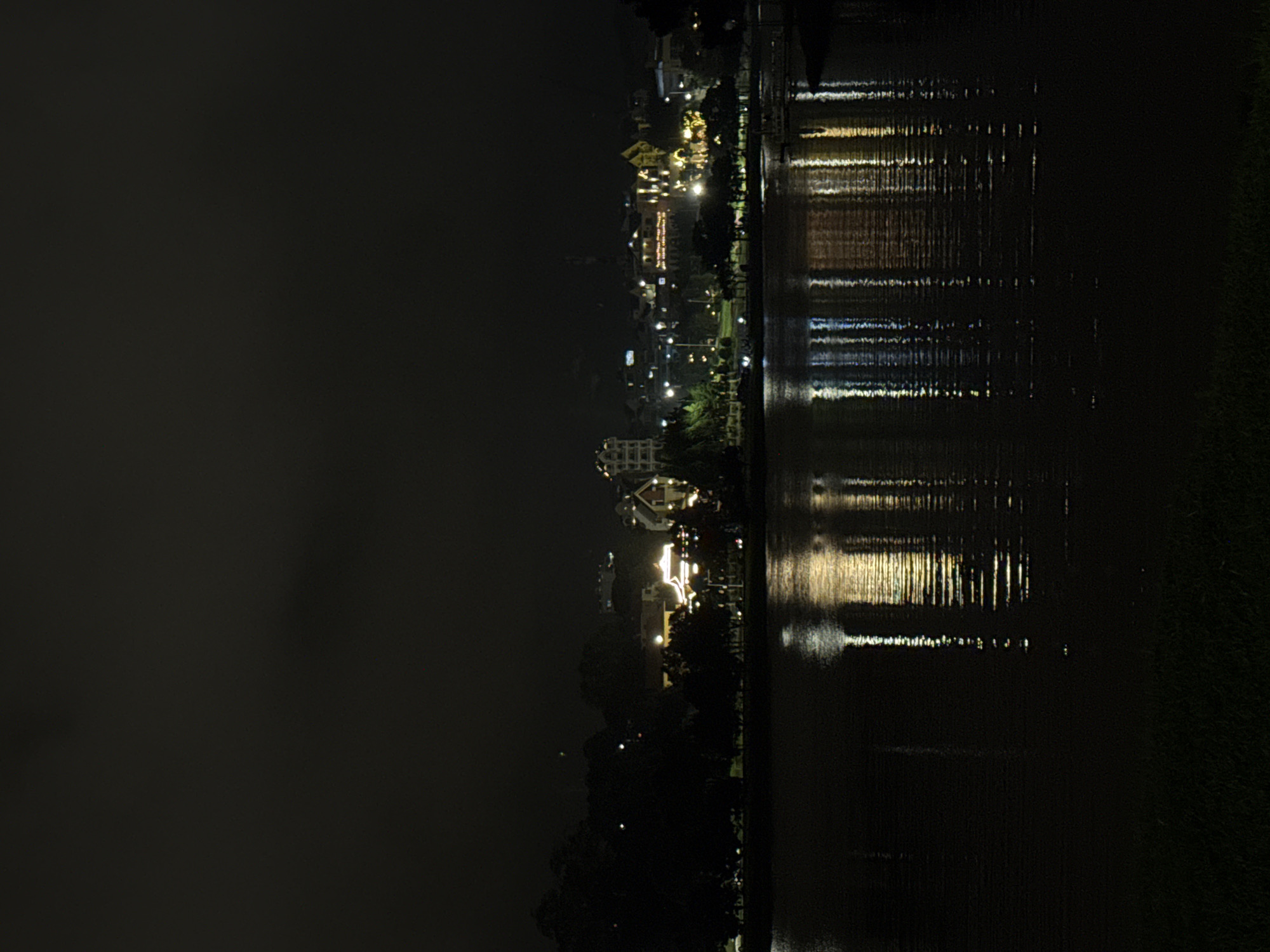 The image captures a serene nighttime view across a body of water, likely a lake or river, towards the illuminated opposite bank in Da Lat, Vietnam. The sky is dark, characteristic of night.
On the far bank, a collection of buildings, possibly hotels or other establishments, are brightly lit with artificial lights, casting warm, inviting glows. Streetlights are visible lining the bank, contributing to the overall illumination. Dark silhouettes of trees are interspersed among the structures and along the waterfront, adding depth to the scene.
The calm surface of the water prominently displays shimmering reflections of all the lights from the opposite bank. These reflections appear as elongated, vertical streaks due to the gentle movement or ripples on the water's surface, creating a dynamic interplay of light and darkness. No specific activities or events are discernible, and no people are visible. The scene evokes a tranquil and picturesque urban night ambiance. There is no visible text in the image.