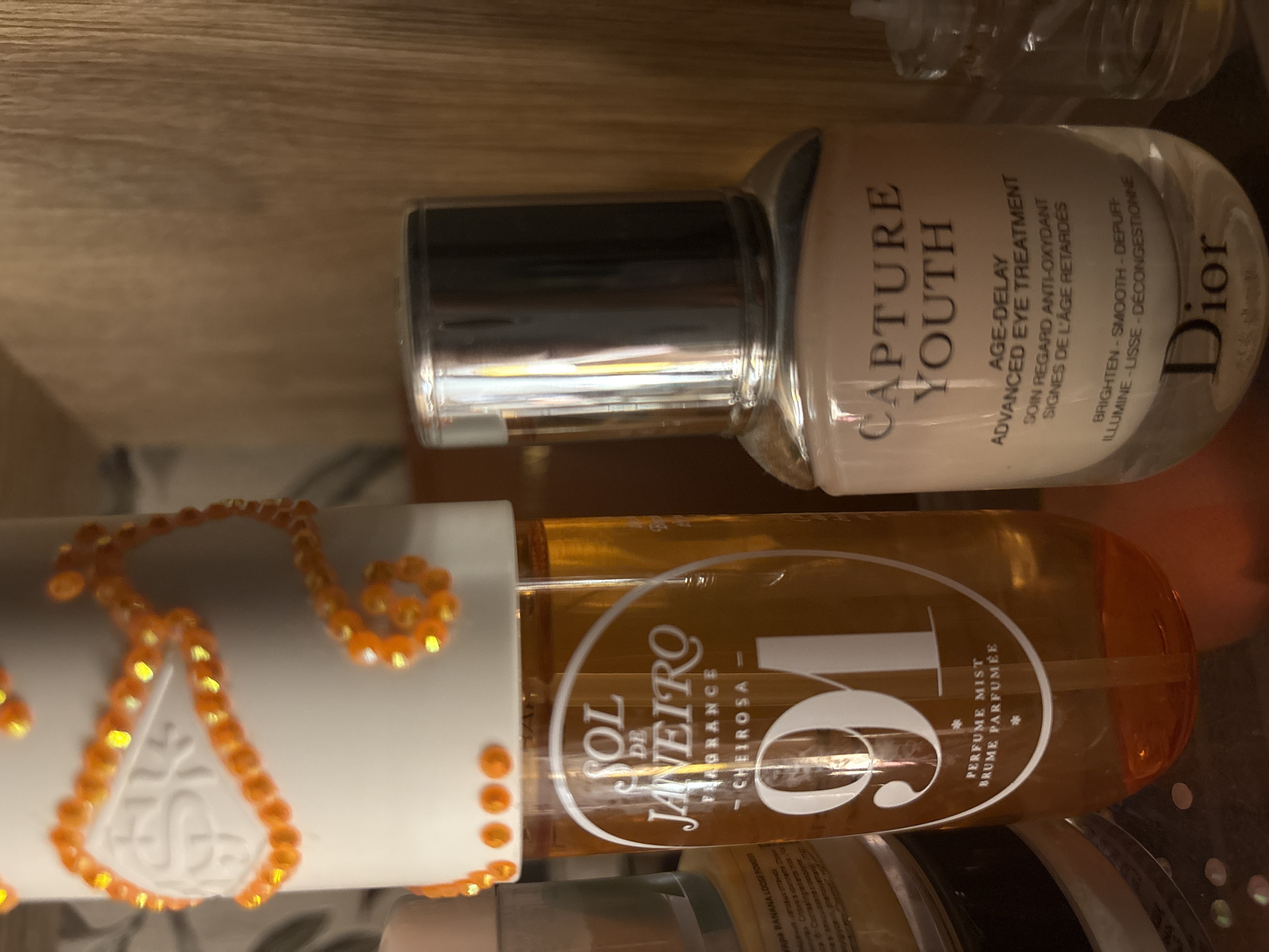 A close-up shot shows two cosmetic bottles on a wooden surface. On the left, a tall, orange-tinted bottle of "SOL DE JANEIRO" perfume mist is decorated with orange beads around its white cap. The label on the bottle reads "SOL DE JANEIRO FRAGRANCE CHEIROSA 91 PERFUME MIST BRUME PARFUMÉE". To the right, a smaller, clear glass bottle of Dior "CAPTURE YOUTH" age-delay advanced eye treatment is visible. Its label reads "CAPTURE YOUTH AGE-DELAY ADVANCED EYE TREATMENT SOIN REGARD ANTI-OXYDANT SIGNES DE L'AGE RETARDÉS BRIGHTEN - SMOOTH - DEPUFF ILLUMINE - LISSE - DÉCONGESTIONNE" and prominently features the "Dior" logo at the bottom. The background is a blurred wooden texture. The scene is indoors. The location is not specified in the image or the provided hints.