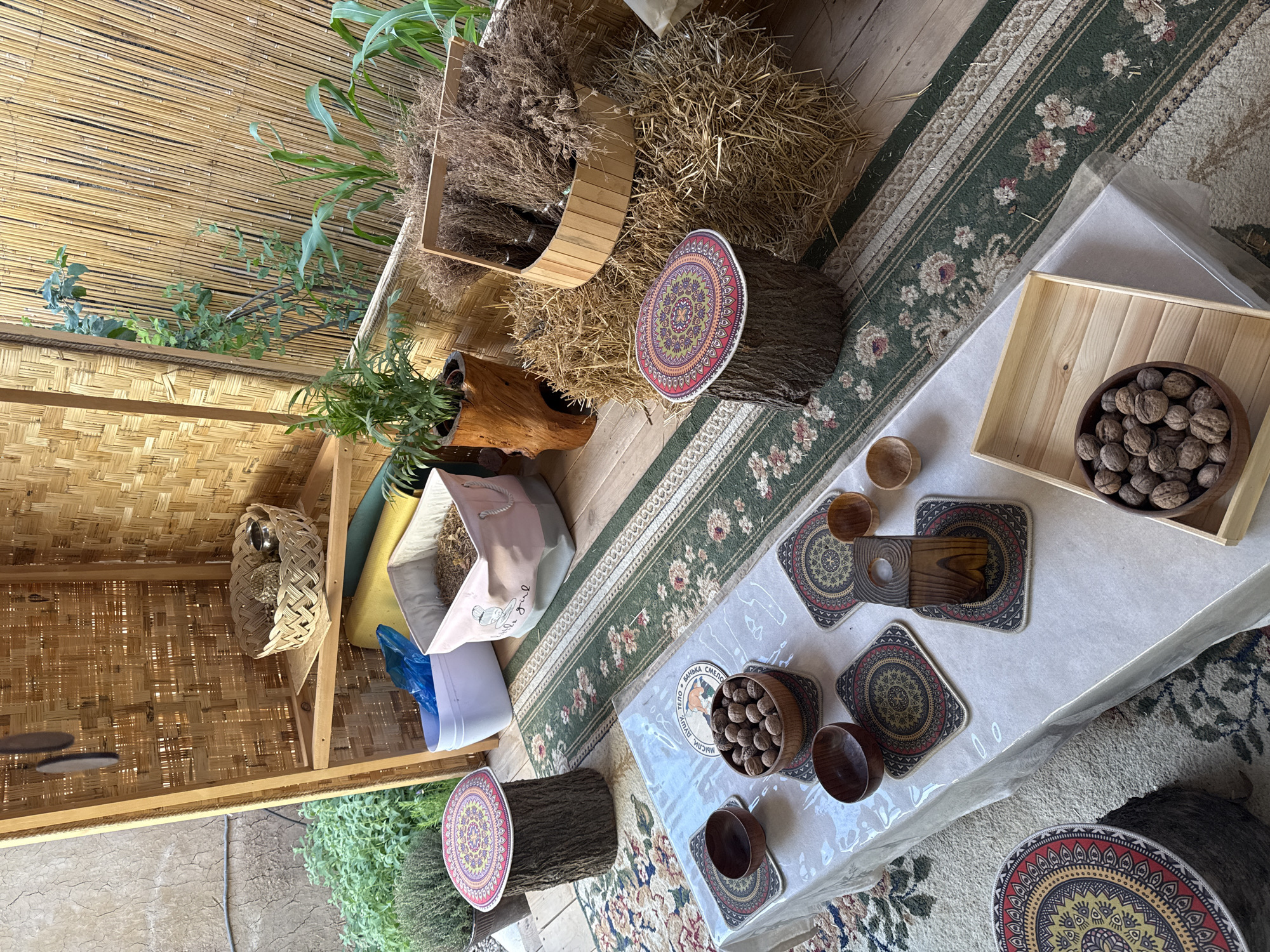 The image shows an outdoor scene in Tashkent, Uzbekistan. The setting appears to be a covered patio or an outdoor display area. There is a wooden structure with shelves on the left holding various items, including baskets, plants, cushions, and containers. The floor is covered with a rug featuring floral patterns and green stripes.
A low table covered with a translucent plastic tablecloth is positioned to the right of the wooden structure. The table is set with wooden bowls, coasters with geometric patterns, and a wooden tray holding walnuts. There are also wooden coasters and tools.
Several tree stumps with decorative circular coverings are used as seating or display surfaces. Hay or dried grass is arranged in baskets and around the stumps. The background features greenery and glimpses of the surrounding environment. No people are visible.