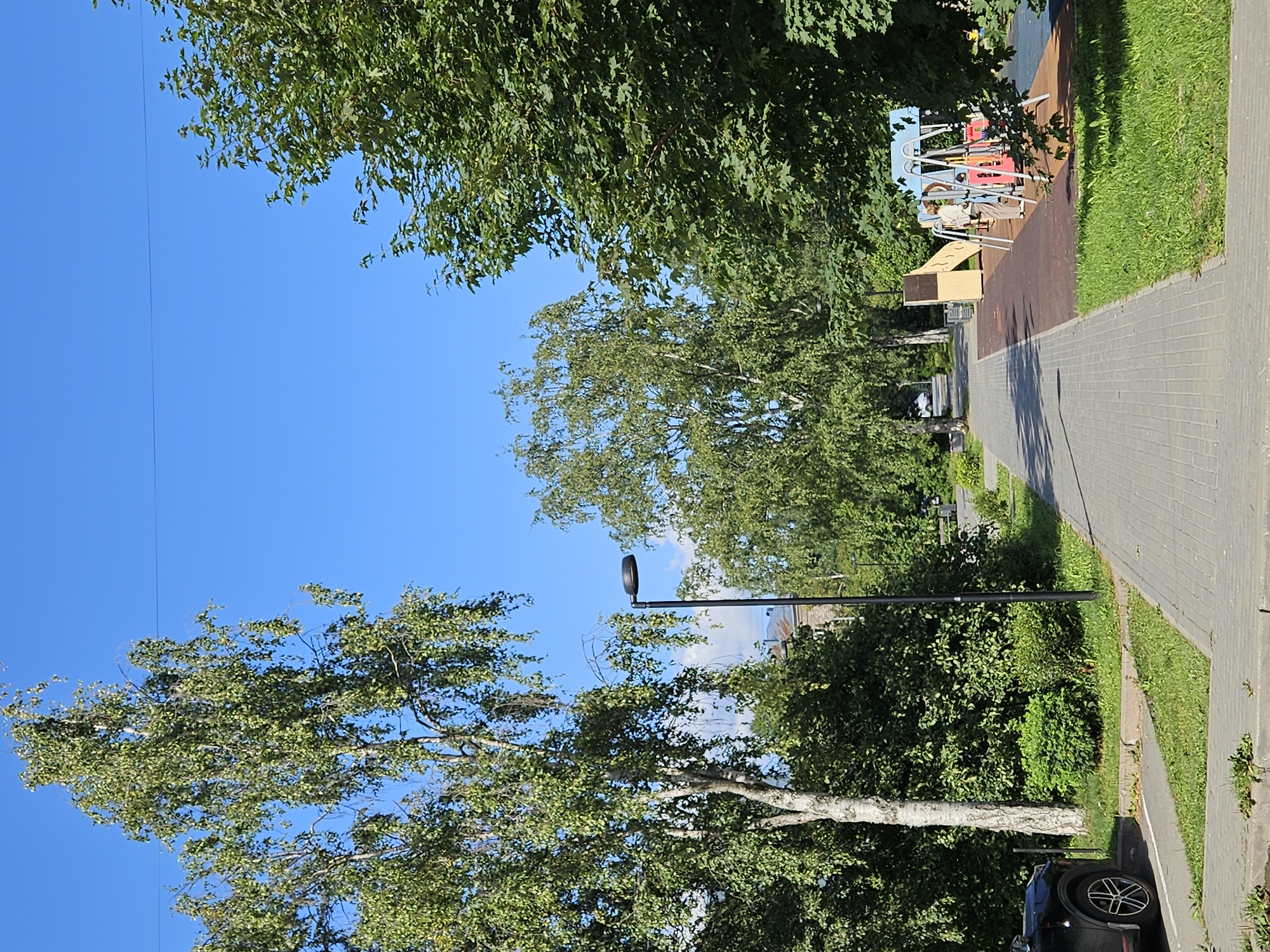 The image shows a paved pathway with a playground to the right and a black car parked to the left. Tall, leafy trees line the pathway, with a clear blue sky overhead. The playground features swings, a slide, and other play structures. A person is standing near the swings, looking at the playground. The scene appears to be in a residential area or park.