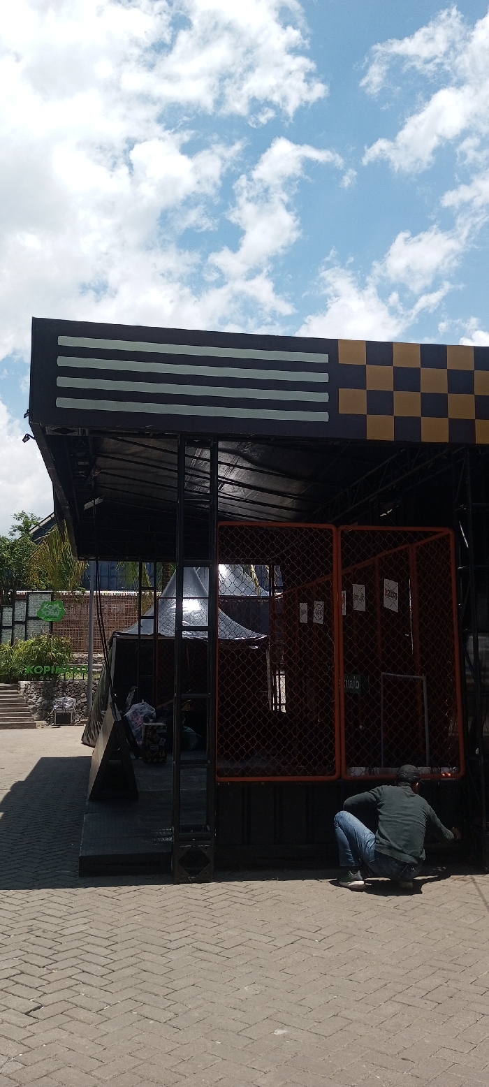 The image displays an outdoor scene in Tagtse Tashidingkha, Bhutan, under a clear blue sky with scattered white cumulus clouds. The ground consists of light grey rectangular bricks laid in a herringbone pattern.
A dark-colored booth-like structure occupies the central and right portions of the frame. Its flat canopy features a black fascia decorated with several horizontal light grey stripes on the left and a black and mustard-yellow checkerboard pattern on the right. The structure's right side is enclosed by two vertical orange mesh panels with a square grid pattern, behind which several white papers are taped to an interior surface. A black tarp is draped over parts of the structure, and a reflective surface is visible within its interior. On the far left of the booth, a dark, angled panel rests against its base.
One individual is present in the lower right, seen from behind, in a crouched posture. The person wears a dark long-sleeved shirt, blue jeans, and a dark baseball cap, and is observed reaching towards the lower, dark-colored base of the booth with their right arm.
In the mid-ground to the left, behind the booth, green foliage and steps leading upwards are visible. A partially obscured green sign in the background reads "KOPI..." and "OBA". The scene is illuminated by direct sunlight, casting shadows.