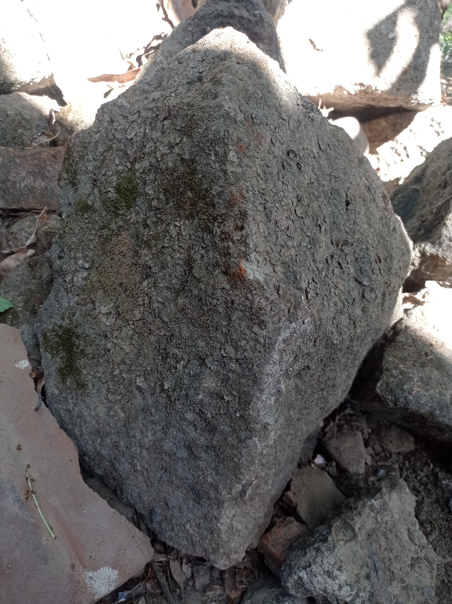The image shows a close-up of several large, rough-textured rocks. The largest rock is in the center, with a mottled gray and brown surface that appears to be covered in a dry, flaky layer and some patches of green moss. Sunlight illuminates the top and right side of this central rock, creating bright highlights and casting shadows. Other rocks are visible surrounding the central one, some appearing darker and smoother. The ground around the rocks is covered with loose dirt, small stones, and dried leaves. A few pieces of what look like broken terracotta or tile are also present, particularly in the lower left and center-right of the image. The overall environment appears to be outdoors, possibly a natural area or a construction site, in Mimbo, Indonesia. There are no people or animals visible in the image.