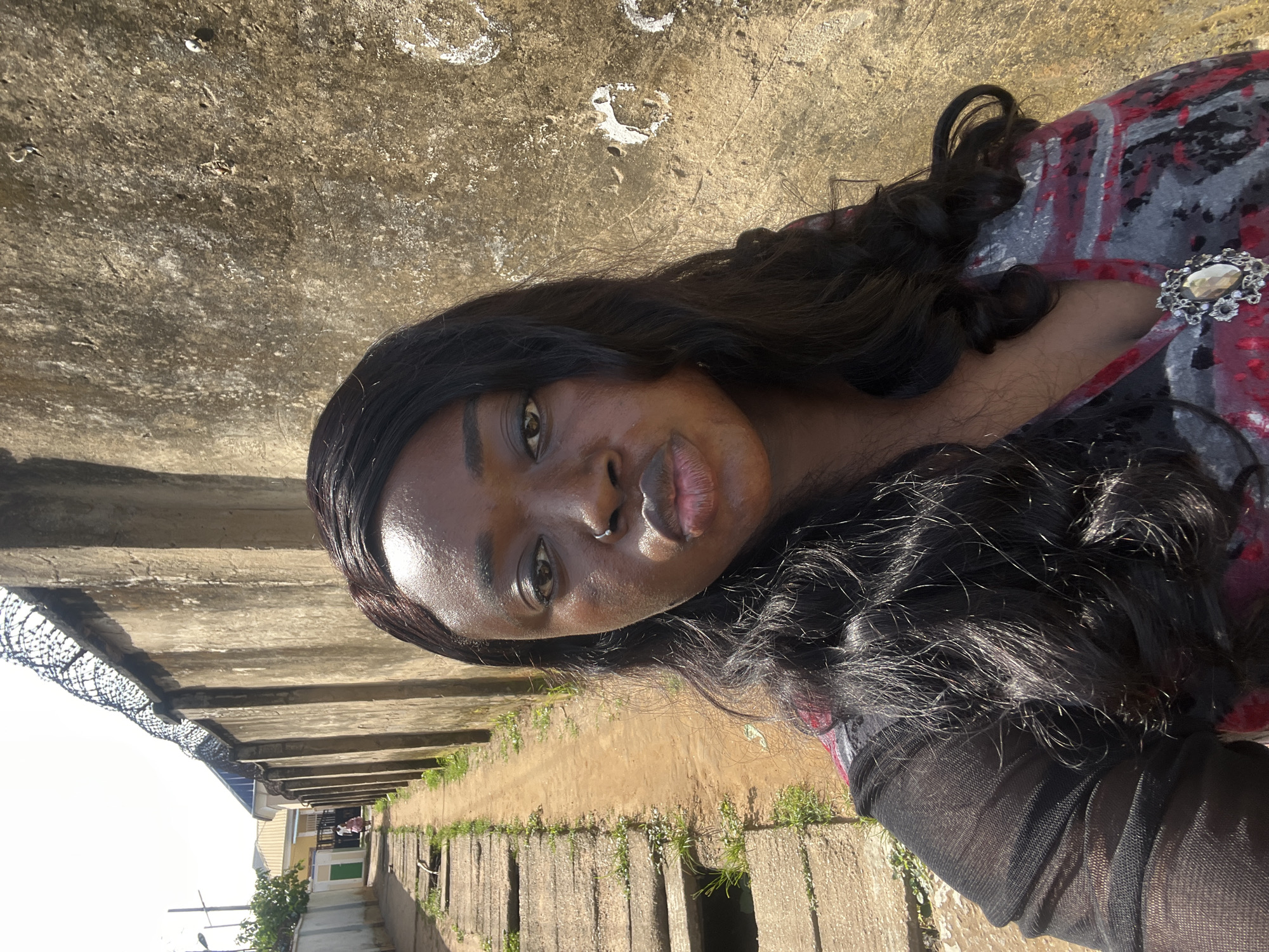A young woman with dark skin and long, dark, wavy hair is standing on a dirt path next to a concrete drainage ditch and a tall, rough-textured concrete wall topped with barbed wire. The woman is wearing a black and red patterned garment and has a silver-toned brooch with an oval gemstone on her chest. Her head is turned slightly towards the camera, and she is making a puckered expression with her lips. The background shows a narrow street with more concrete structures and some vegetation. The scene is illuminated by bright daylight, suggesting an outdoor setting likely within a residential or less developed area of Abuja, Nigeria.