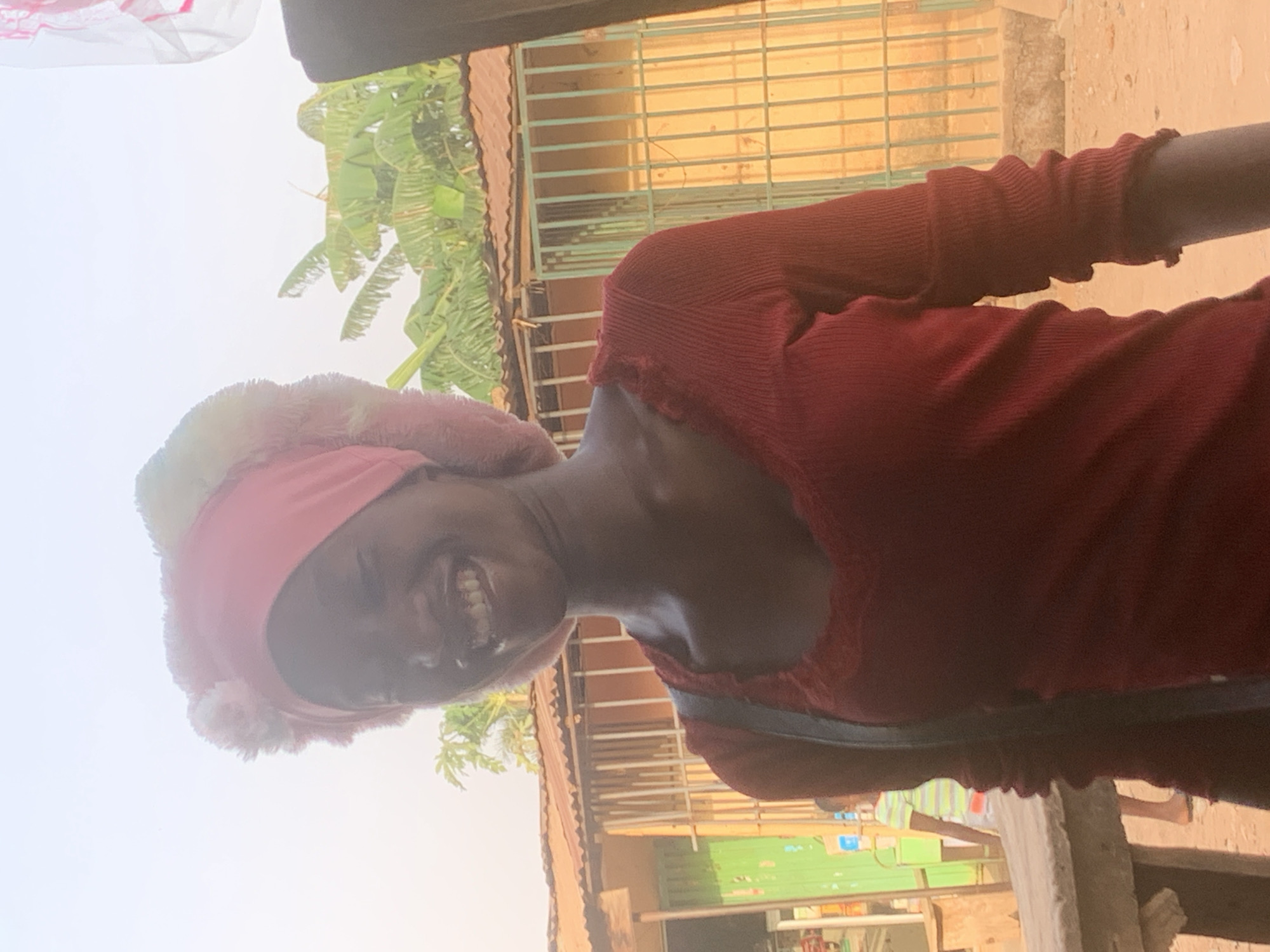 A woman wearing a red long-sleeved top and a pink headscarf is smiling at the camera. The background includes a building with a red corrugated iron roof and a light green wall. A banana plant is also visible, and there is another person visible at a distance. The location is potentially in Sabon Tasha, Nigeria.