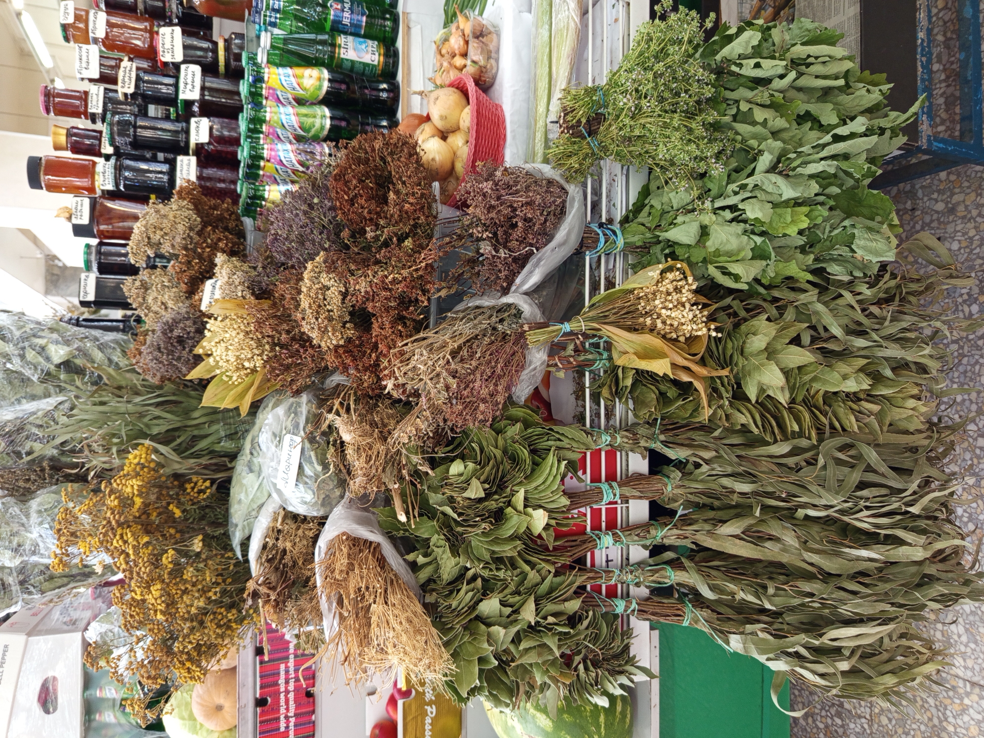 The image depicts a stall at an outdoor market, likely in Saint Petersburg, Russia, selling a variety of dried herbs, plants, and foodstuffs. The foreground is dominated by hanging bunches of dried herbs, primarily large leafy varieties in shades of green and brown, some tied with blue and green twine. To the left, a large green watermelon is partially visible. Behind these hangings, a display of dried floral arrangements and plants is evident, featuring yellow flowers, brown clusters, and long, straw-like stalks. In the mid-ground and background, shelves are laden with jars of preserved goods, likely jams or honey, with labels in Cyrillic script. Bottles of beverages, some appearing to be sodas, are also present. A basket of onions sits on a shelf. The overall impression is a vibrant and abundant market scene, showcasing natural products and local produce. The lighting suggests it is daytime.