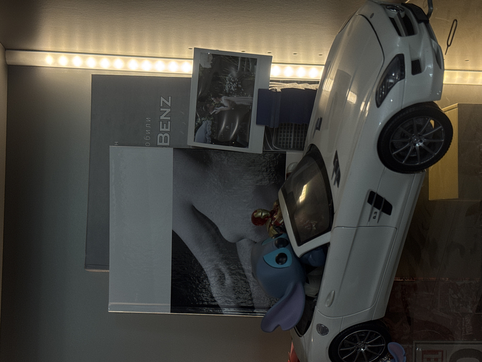 The image shows a shelf with a white toy car, a "Stitch" figurine, a book titled "BENZ", a photograph, and other objects. The lighting suggests an indoor environment. No evidence suggests the location is in Khimki, Russia.