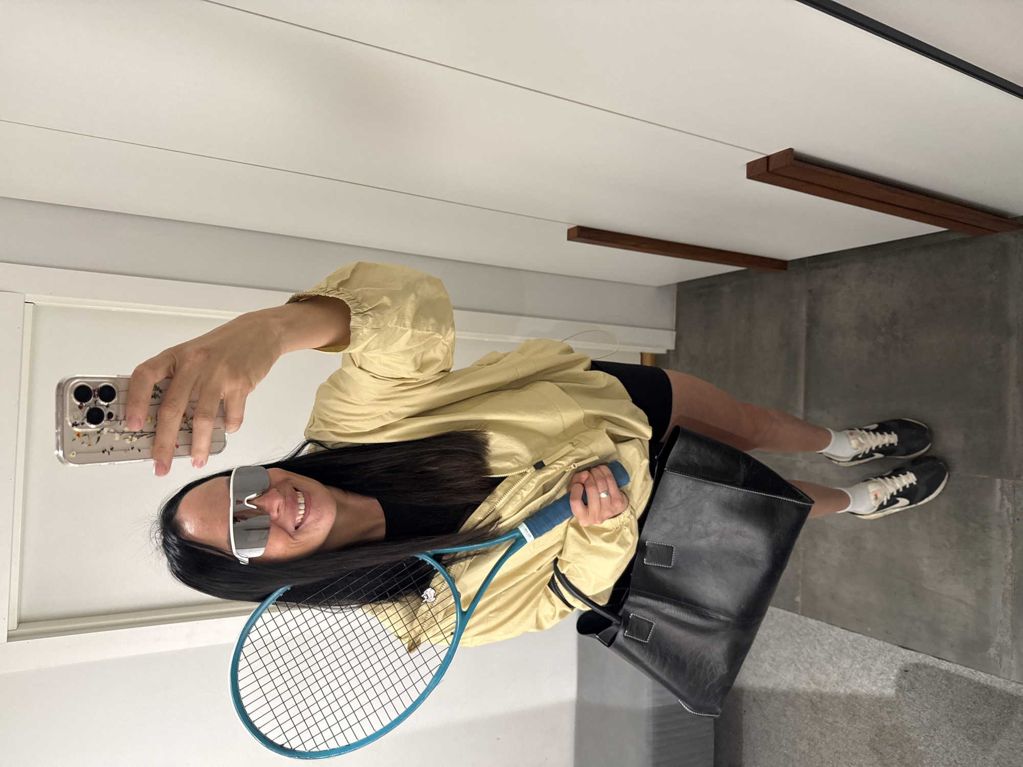 A woman stands in a room, taking a mirror selfie. She is holding a turquoise tennis racket in her left hand and a large black leather tote bag in her right. She is wearing reflective sunglasses, a light yellow jacket, black shorts, white socks, and black and white athletic shoes. The background shows a white wall with vertical wooden accents and a door. A gray upholstered bench is visible to the left. The floor is a concrete-like material. The woman's phone, with a floral case, is visible in her hand as she takes the photo. The image does not provide any specific details about the city of Chaikovsky, Russia.