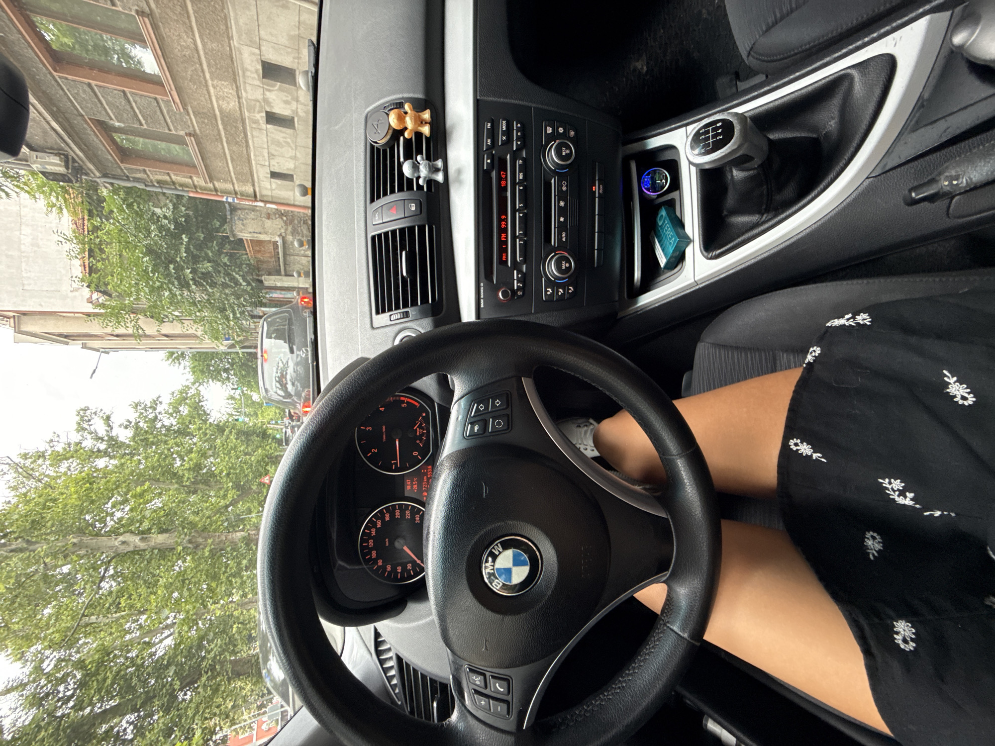 The image shows the interior of a BMW car, viewed from the driver's perspective. The steering wheel, dashboard with gauges, central console with a radio and climate controls, and gear shifter are visible. The driver's legs are visible, wearing a black dress with white floral embroidery. Outside the car, there is a street scene with trees, buildings, and other cars, suggesting the vehicle is in traffic. The buildings are typical of European architecture, with some featuring ornate windows and aged facades. The overall impression is of a person driving through a moderately dense urban environment, likely in Plovdiv, Bulgaria, given the prompt's context. There are two small figurine air fresheners on the dashboard.