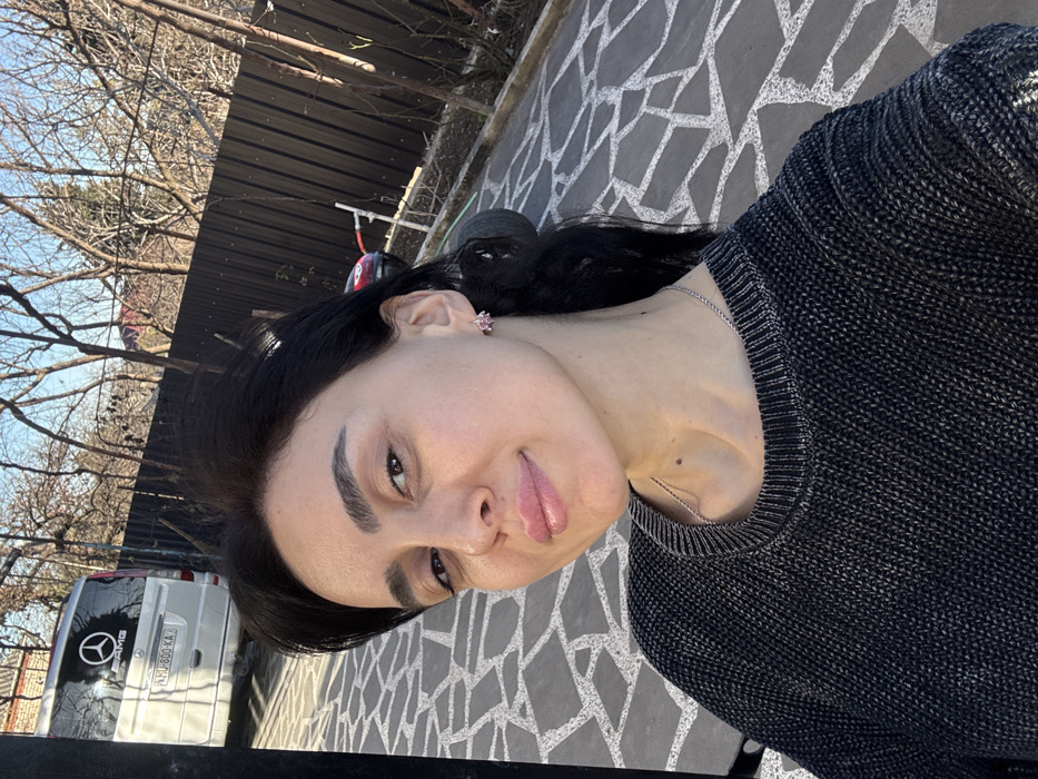 The image presents a close-up outdoor portrait of an adult female situated in Bolnisi, Georgia. The woman, positioned centrally, has dark hair, a light complexion, and a slight smile while looking directly at the camera. She is attired in a dark-colored, possibly charcoal, knit sweater with a ribbed collar, and a thin silver-toned chain necklace. A small, pink, flower-shaped earring is visible on her right earlobe. The immediate environment consists of a paved surface made of irregularly shaped light-colored stone tiles. In the background to the left, a white Mercedes-Benz Sprinter van is parked on an incline, featuring a black "AMG" logo on its rear window and a visible Georgian license plate "UU-800-KA". To the right, a dark, horizontal-slatted wooden fence extends across the mid-ground. Beyond the fence, several bare trees are visible against a clear blue sky. Direct sunlight illuminates the scene, casting shadows on the paved ground. A red object is partially discernible behind the fence on the far right.