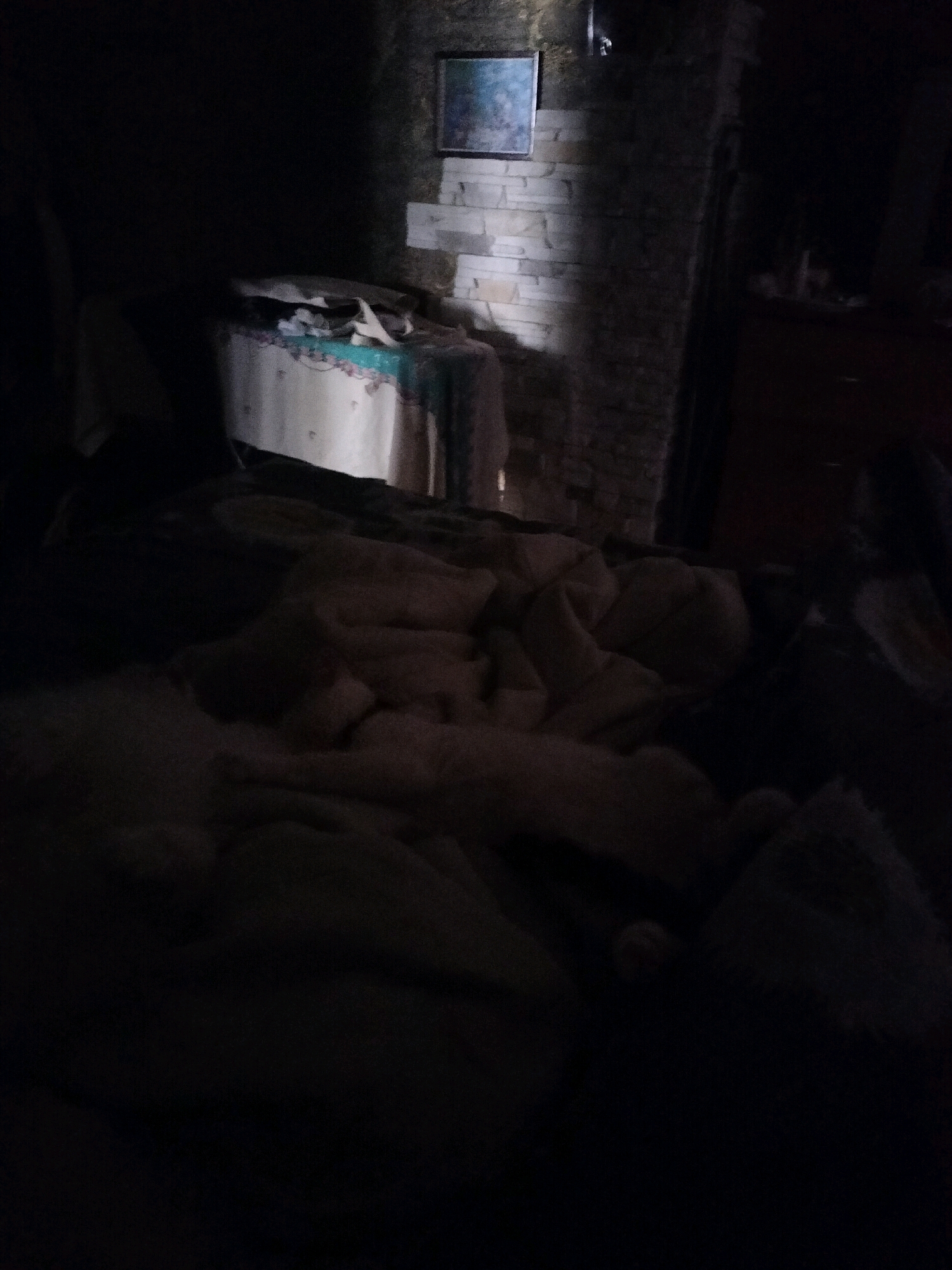 The image depicts a dimly lit interior scene. In the background, a fireplace with stacked stone is visible, adorned with a framed picture at the top. To the left of the fireplace, a table covered with a patterned cloth holds various items, including what appears to be fabric. In the foreground, a bed is covered with blankets, creating a rumpled appearance. The lighting suggests it is either nighttime or the room is enclosed and lacking natural light. The context of Zhytomyr, Ukraine, is not discernible from the visual elements of the image.