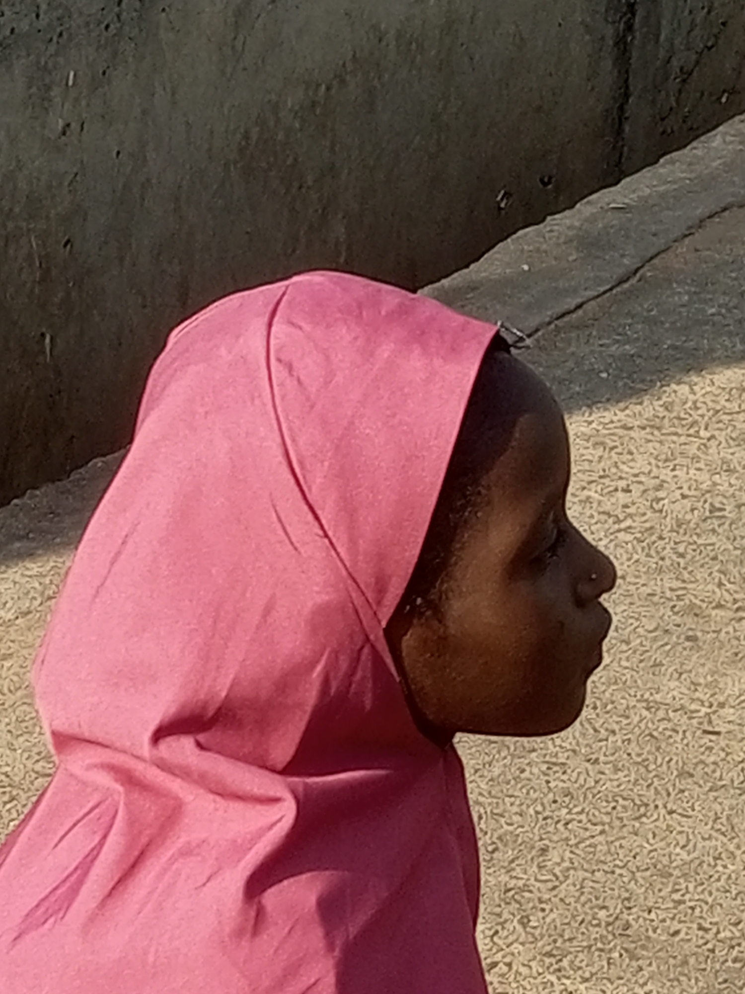 This is a medium shot of a young Black girl from the side, wearing a pink head covering. She is looking to the right with her mouth slightly open. The background consists of a textured, light-colored surface and a darker, rough-textured wall behind that. The lighting suggests it is daytime, likely with direct sunlight casting shadows. The scene appears to be outdoors in an urban or semi-urban environment, possibly a street or courtyard. There is no visible text in the image.