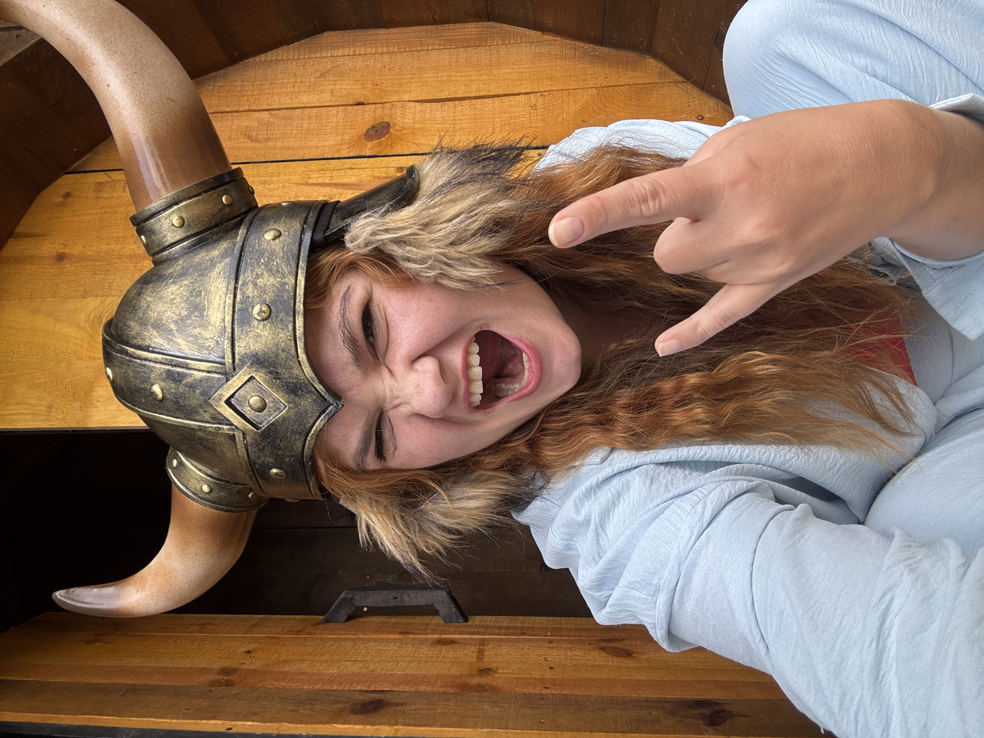 A close-up, eye-level, medium shot captures a young woman with long, wavy red hair, wearing a golden-bronze Viking helmet with large brown horns and furry ear flaps. She is wearing a light blue, textured hooded garment. Her mouth is wide open in a fierce expression, and her right hand is raised in a rock and roll gesture. The background consists of a wooden structure, possibly a doorway or wall, with vertical planks and a dark opening to the left. The lighting is natural and appears to be from the front, illuminating the woman's face and attire. The setting does not provide specific geographic indicators of Thôn 1, Vietnam, and the scene appears to be a staged or costume-related photograph.