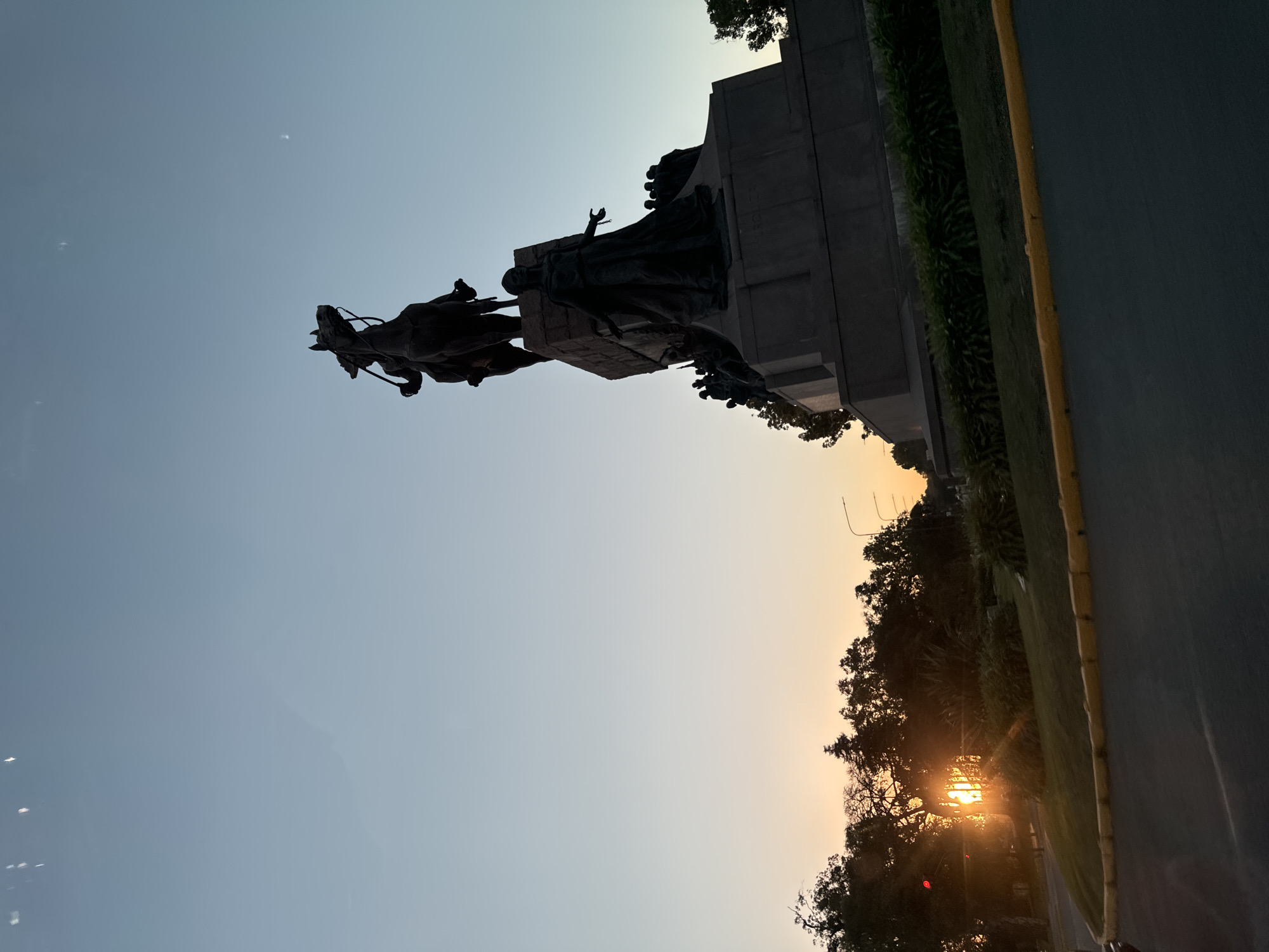 The image captures a bronze equestrian statue set against a sunset sky. The statue depicts a man on horseback, with additional figures surrounding the base of the monument. The monument is located alongside a road and some trees. The location is Buenos Aires, Argentina.