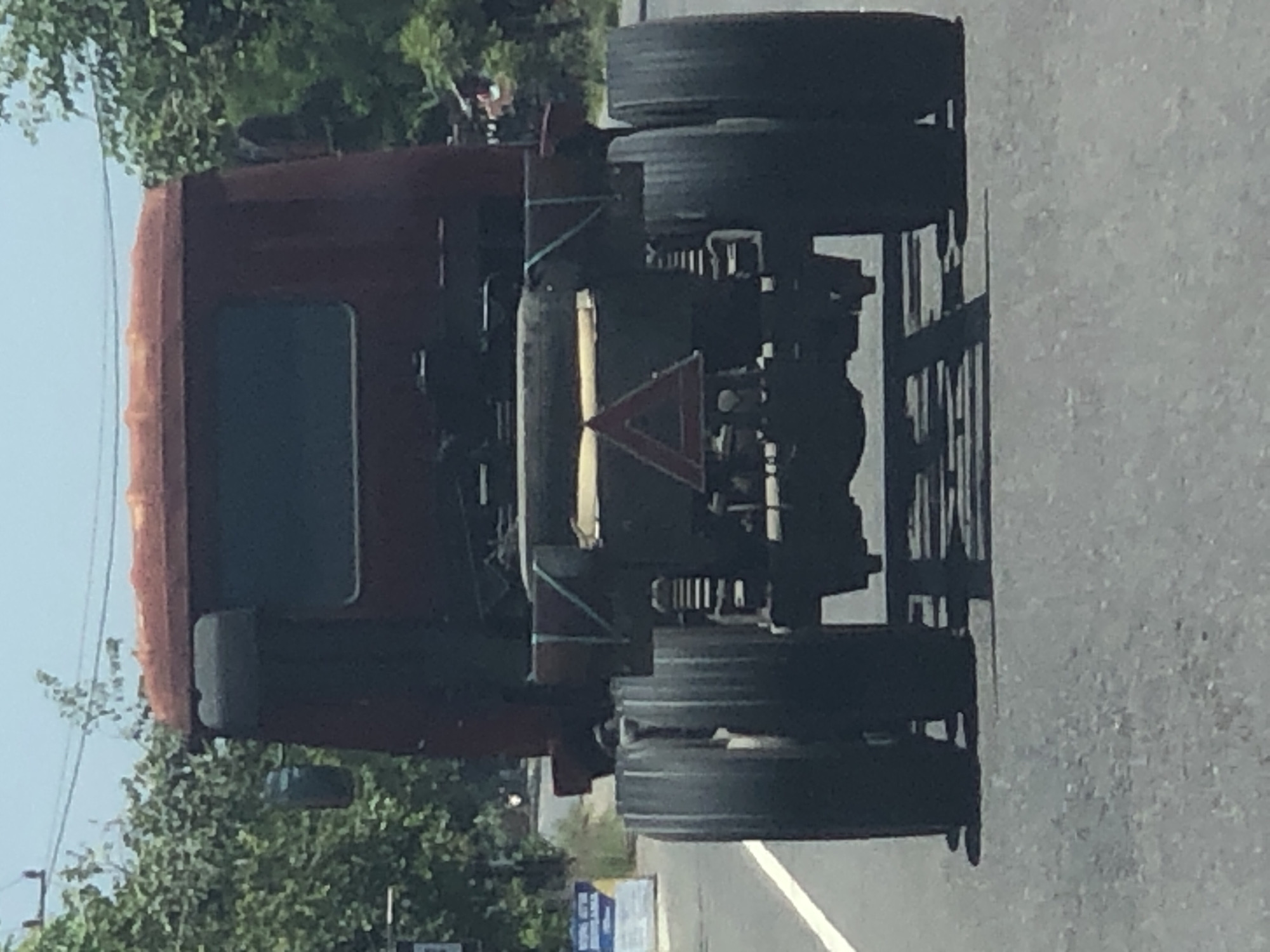The image shows the rear view of a red semi-truck driving on a paved road. The truck has dual rear wheels on each side. A red triangular sign with a white border and a white symbol inside is attached to the rear chassis of the truck. The truck is on a road with a white line marking the left lane. Trees and foliage are visible on either side of the road, and the sky is clear and bright. A sign with blue and white text is visible on the left side of the image, in the background. The scene appears to be set in Matangneuheun, Indonesia, based on the provided context, likely during daylight hours given the bright lighting conditions and shadows. There are no people or animals visible in the frame.