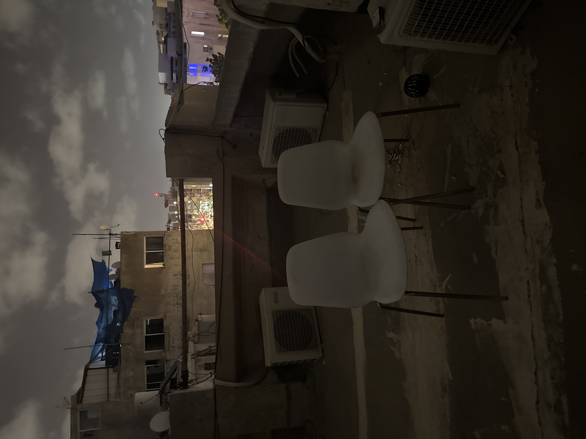 The image captures a dimly lit rooftop scene in Tel-Aviv, Israel at night. Two white chairs are centrally positioned, surrounded by air conditioning units mounted on adjacent walls. The urban environment is visible in the background, featuring buildings and cloudy skies. A partial blue tarp is attached to a nearby building. Wires and satellite dishes are also visible.