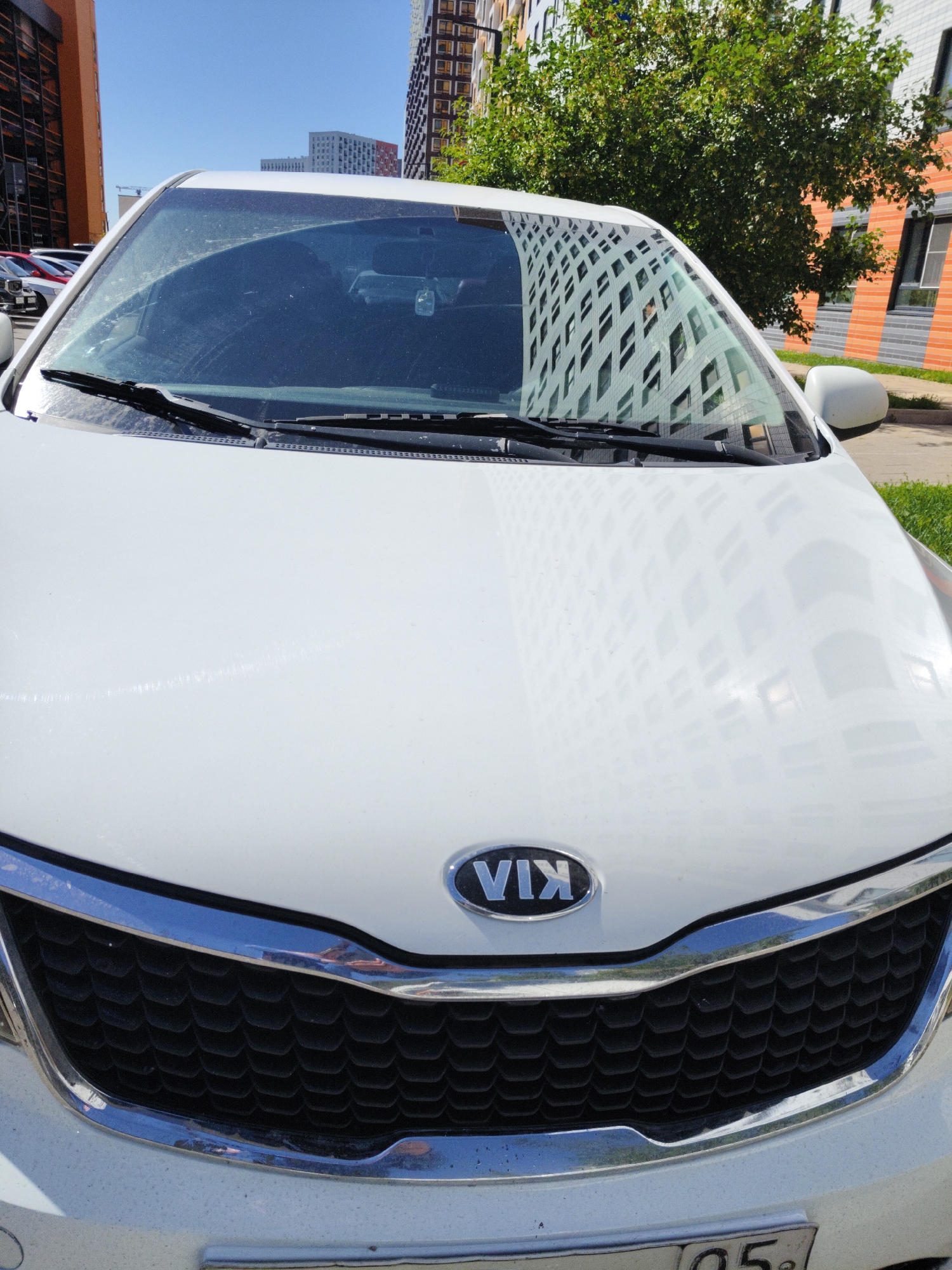 A white Kia car is parked in an urban environment. The car's front is visible, including its grille with the Kia logo. The windshield reflects a modern apartment building with a grid-like facade and trees. The background shows tall apartment buildings and a clear blue sky. The scene appears to be in the city of Kommunarka, Russia, based on the license plate which includes Cyrillic characters and numbers that are consistent with Russian plates. There are no people or animals visible in the image, and no actions or interactions are taking place. The car is stationary.