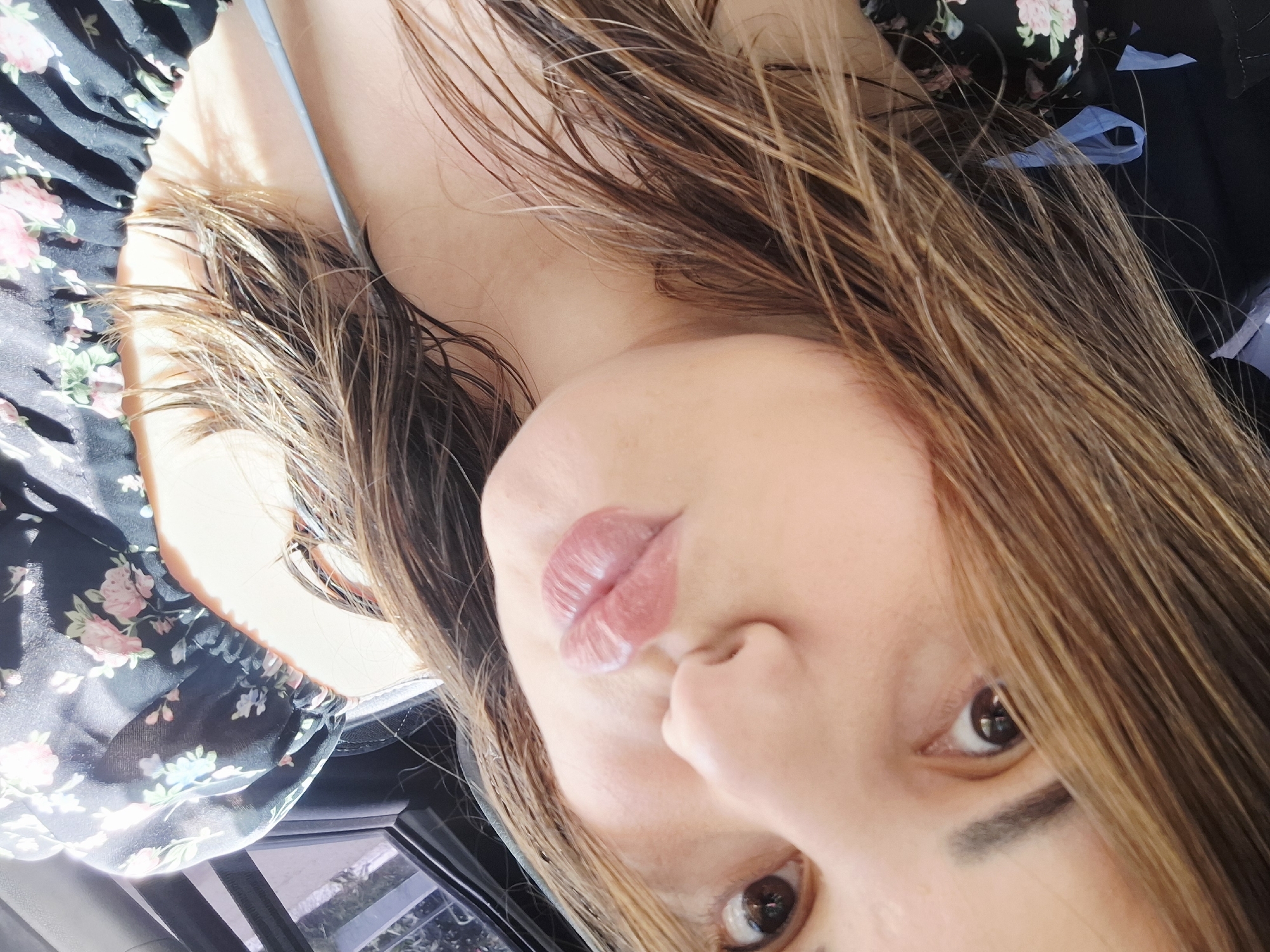 A close-up, selfie-style photo shows a woman's face, from the nose down, as she pouts her lips. Her hair, a medium brown with highlights, is damp and styled loosely around her face. She is wearing a black top with a floral print of pink and blue flowers. The background suggests she is in a vehicle, with a portion of a car window visible on the right side of the frame. Sunlight is casting bright highlights on her hair and skin. There is no information to indicate this image is located in Chaikovsky, Russia.