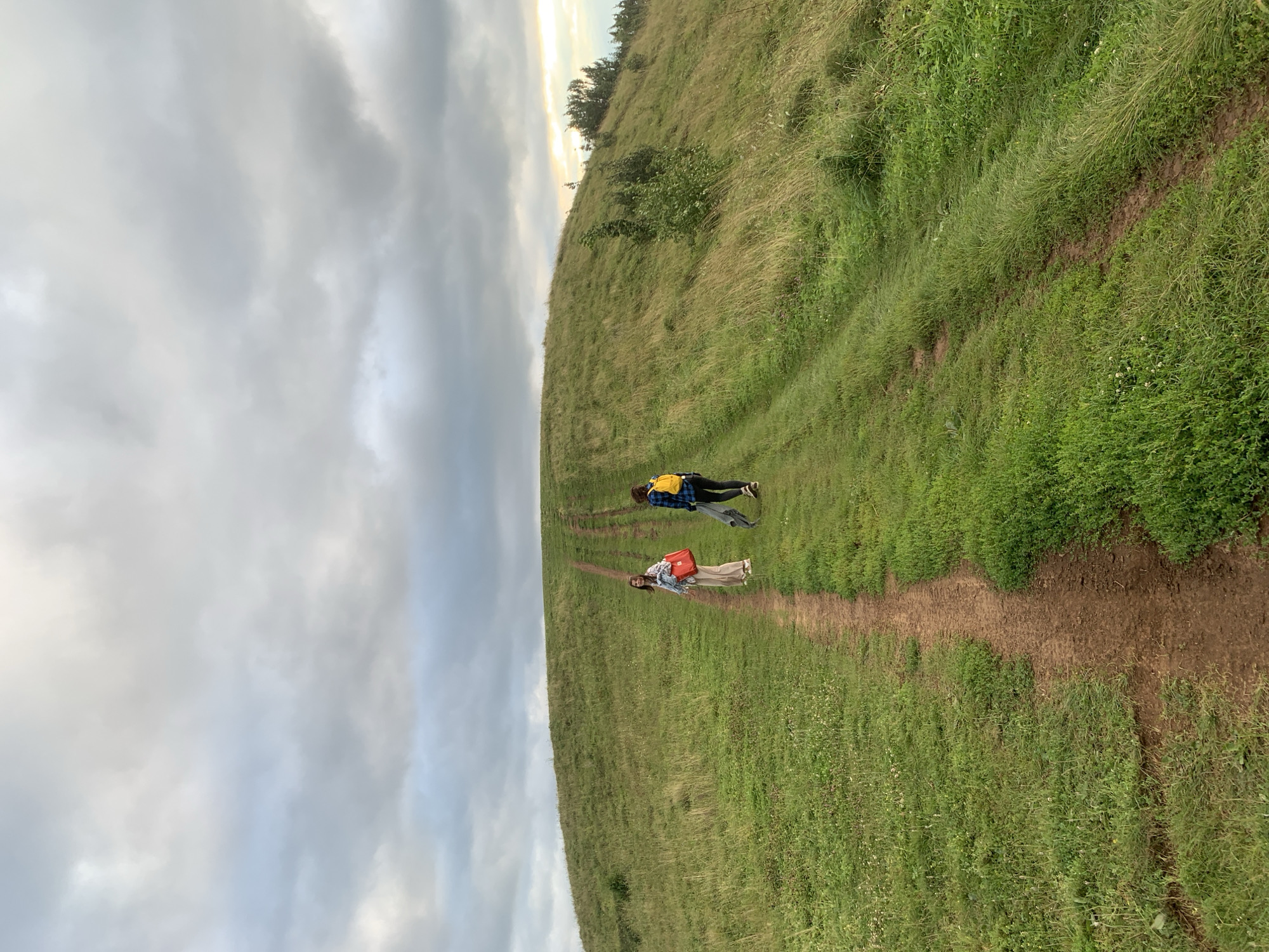 This is a landscape photograph taken in the city of Olkhovka, Russia, on a cloudy day. The scene features two individuals walking up a grassy hill on a dirt path. The person in the foreground, closer to the viewer, is a woman wearing light-colored pants and a white top, carrying a red bag. She is looking back towards the camera. Behind her, further up the path, is another person, possibly a young man, wearing a blue plaid shirt and black pants, with a yellow backpack. This individual is carrying a dark-colored jacket over their shoulder. The hill is covered in lush green grass with some patches of dried grass and small wildflowers. The path itself is a well-worn dirt track that curves upwards. The sky above is filled with heavy, grey clouds, with a hint of lighter sky and possibly sunlight breaking through on the far right side of the image. In the distance, the terrain rolls into more grassy hills.