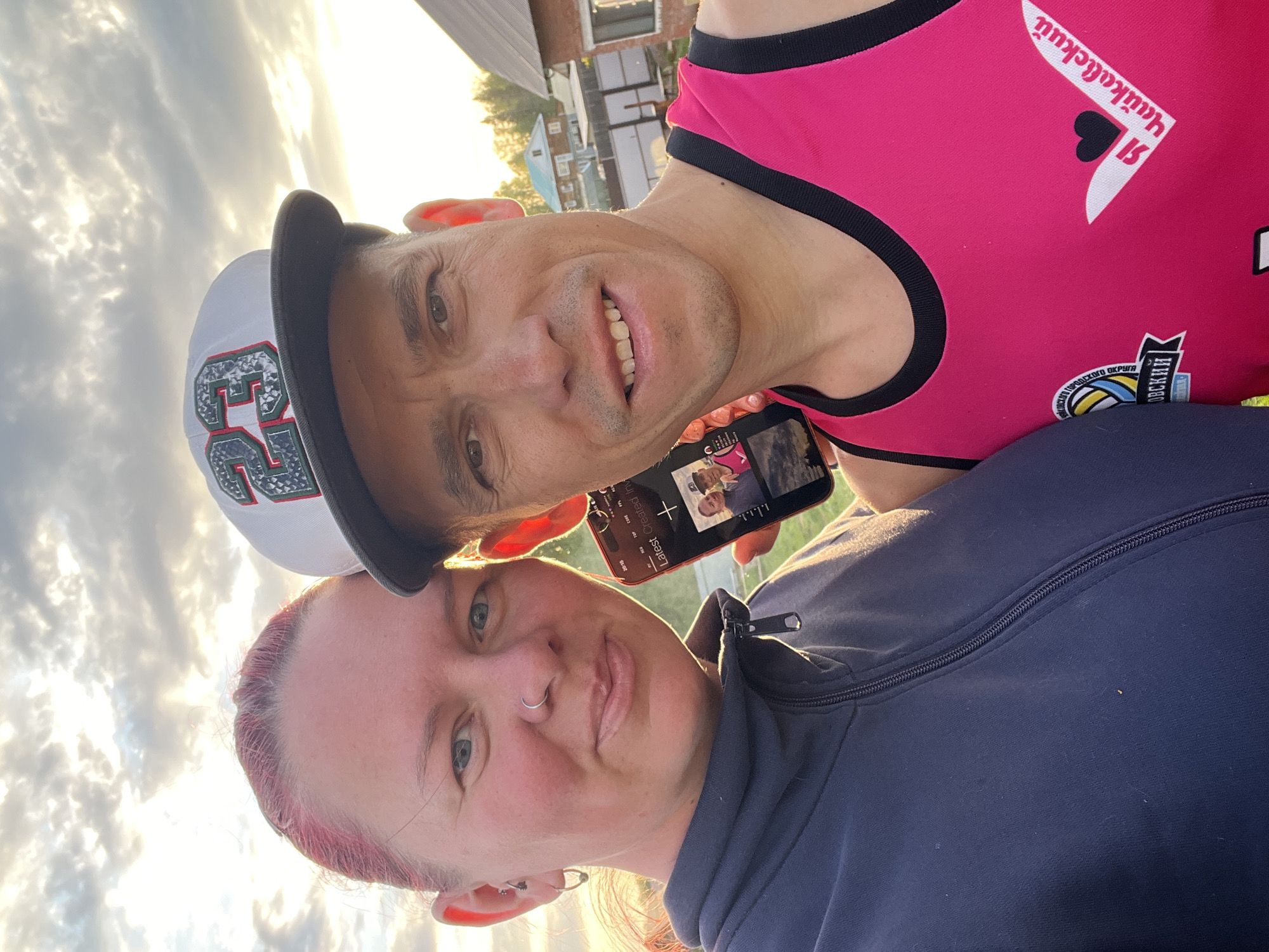 A selfie photograph features two individuals against a backdrop of a sky filled with clouds and a hint of sunset. The photograph appears to have been taken in Chaikovsky, Russia, given the Cyrillic text on the man's shirt. The man is wearing a white and black baseball cap with the number "23" on it, along with a pink tank top that has Cyrillic writing and a heart symbol on it. He is holding a phone in front of him, displaying what looks like a photo or video on the screen. The woman is wearing a dark blue zipped top. A building is partially visible in the background.