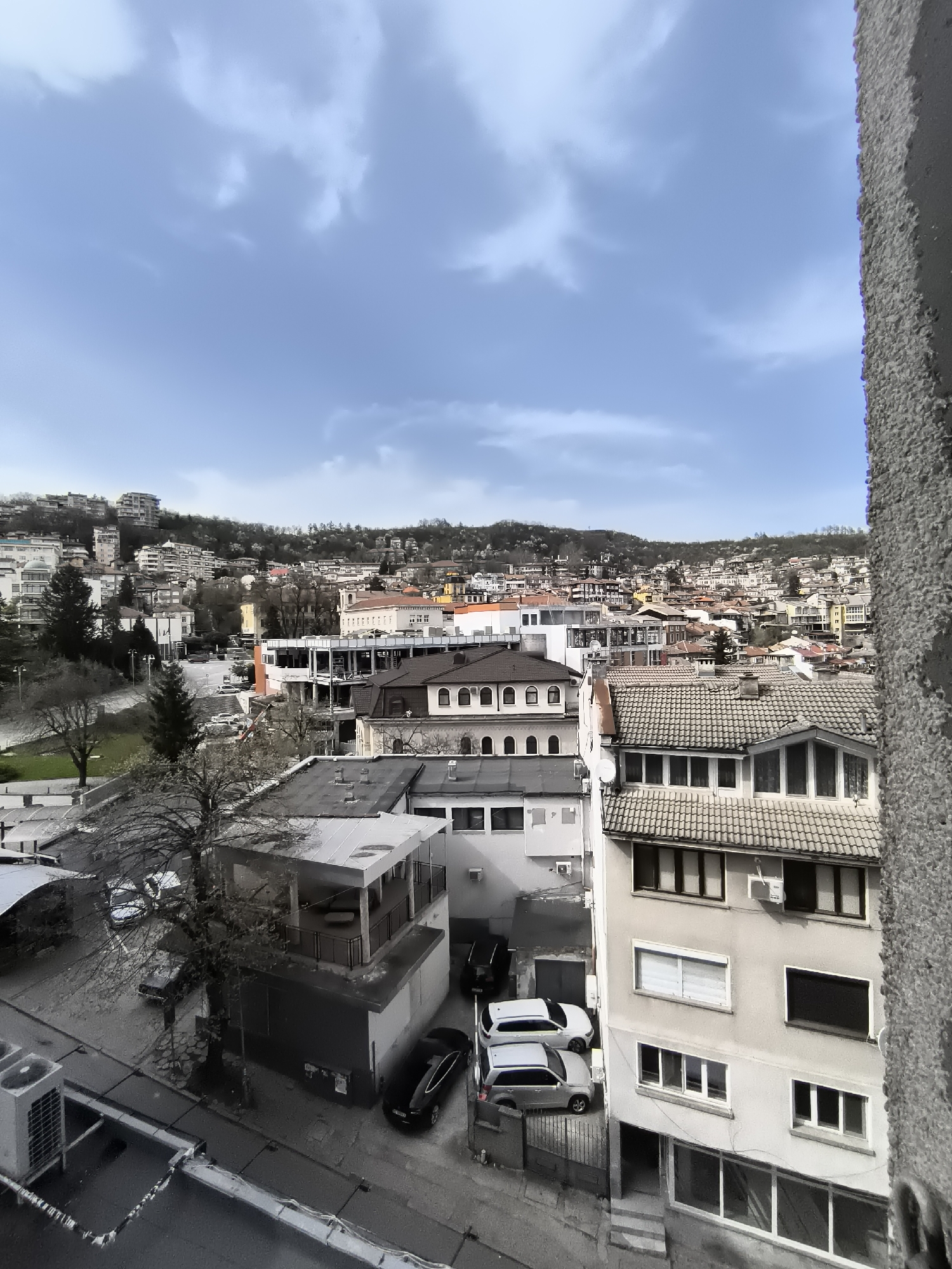 The image shows a cityscape in Veliko Tarnovo, Bulgaria, under a cloudy sky. The buildings are a mix of architectural styles, some with tiled roofs and others with flat roofs. A building under construction is visible in the middle ground. Cars are parked on the street. The buildings are clustered together, giving the impression of a densely populated area. A grey textured object, possibly part of a building facade, is on the right side of the image.