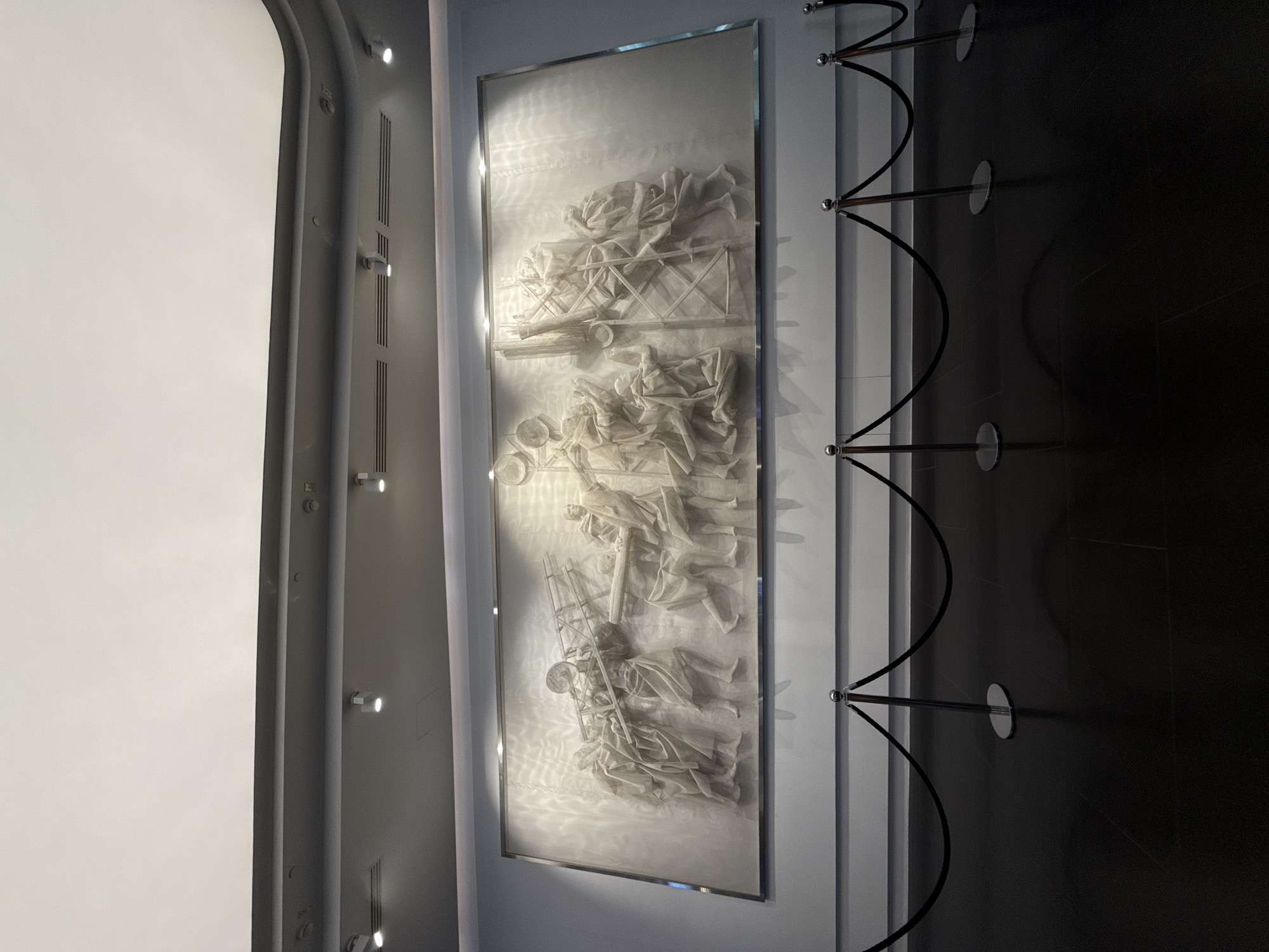 A vertically-oriented, medium shot shows a framed bas-relief art piece hanging on a wall in an interior space. The art piece depicts multiple figures arranged in a layered, sequential manner. The top layer shows figures interacting with a structural element resembling a bridge. The middle layer shows figures in a semi-circular grouping. The bottom layer shows figures with a ladder-like structure. The art piece is framed with a thin metallic silver frame. To the left of the art piece is a light-colored wall with recessed lighting. To the right of the art piece are three black stanchion ropes with silver poles and bases. The scene appears to be in a gallery or museum-like setting. The location is Khimki, Russia. There are no people visible in the image itself. The subjects are the figures depicted in the bas-relief. The objects include the art piece, the frame, the wall, the lights, the stanchion ropes, poles, and bases. The actions are the interactions of the figures within the bas-relief. The environment is an interior space, likely a gallery or museum.