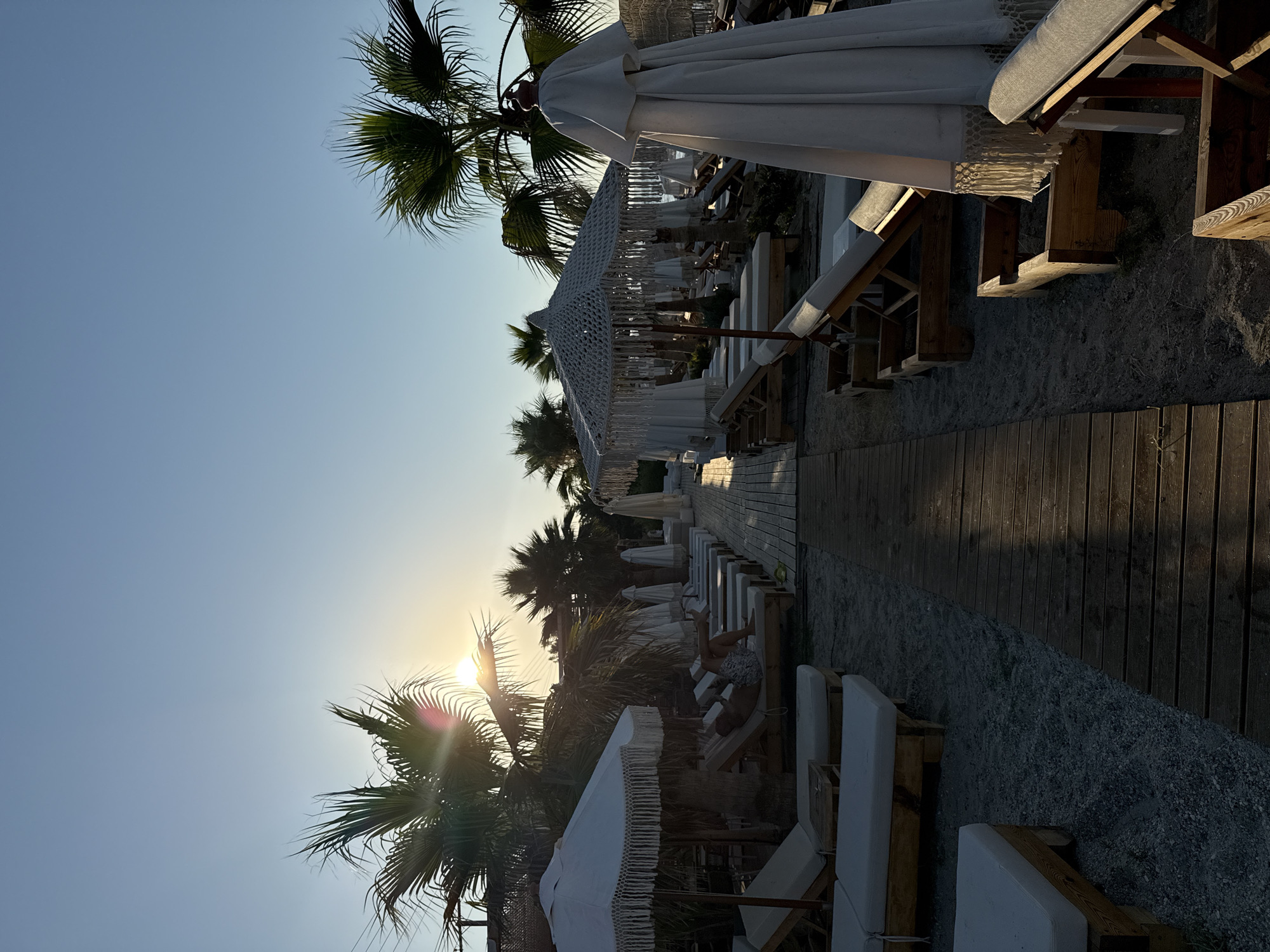 This image depicts a beachside setting in Pallini Municipal Unit, Greece, likely during a warm day as suggested by the clear sky and palm trees. The scene features a row of empty lounge chairs and umbrellas set up on a sandy area, with a wooden pathway running alongside them. The sun is low in the sky, casting long shadows and creating a golden hour glow, indicating it is either sunrise or sunset. The presence of lounge chairs and umbrellas suggests a resort or a public beach area designed for relaxation and sunbathing.
In the foreground, several white umbrellas with decorative fringes are visible, along with wooden-framed lounge chairs topped with white cushions. Beyond these, more lounge chairs and umbrellas stretch into the distance. The beach appears to be relatively quiet, with only one person visible in the distance, lying on a lounge chair. Palm trees are scattered throughout the scene, adding to the tropical or Mediterranean atmosphere. The ground is a mix of sand and small pebbles, with a wooden deck or boardwalk extending along the edge of the beach. The overall mood is serene and peaceful, evoking a sense of vacation and relaxation. No visible text is present in the image.