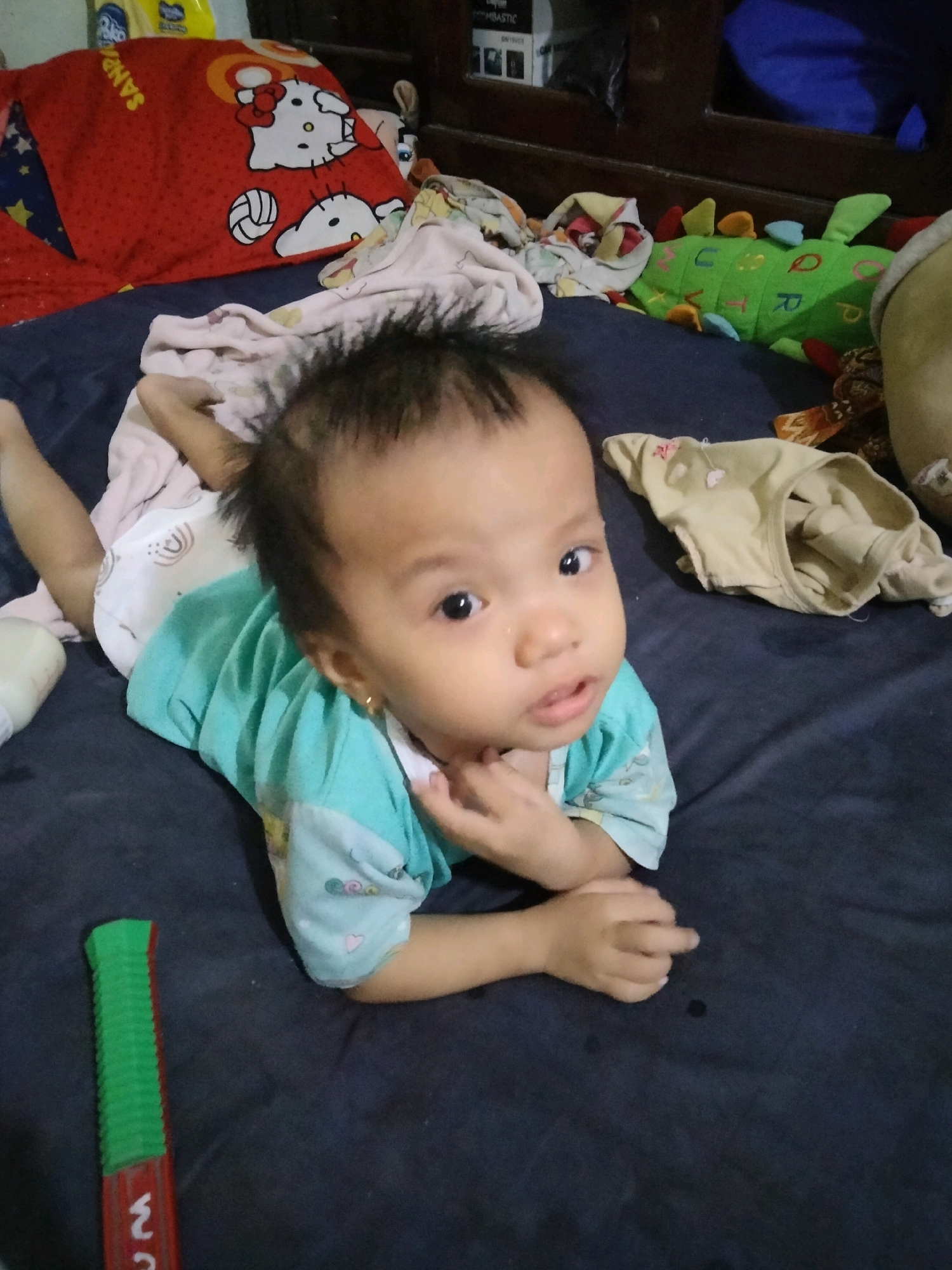 In Suka Sari, Indonesia, a baby is lying on a dark blue surface, possibly a bed or floor, in a dimly lit room. The baby, wearing a turquoise and patterned shirt and a diaper, is positioned on its stomach and looking directly at the camera with a curious expression. Its right hand is near its mouth, and its left arm is bent. To the left of the baby, a green and red toy with some writing is visible. In the background, there are several items including a red pillow with a Hello Kitty design, a green stuffed dinosaur, and other blankets and fabrics.