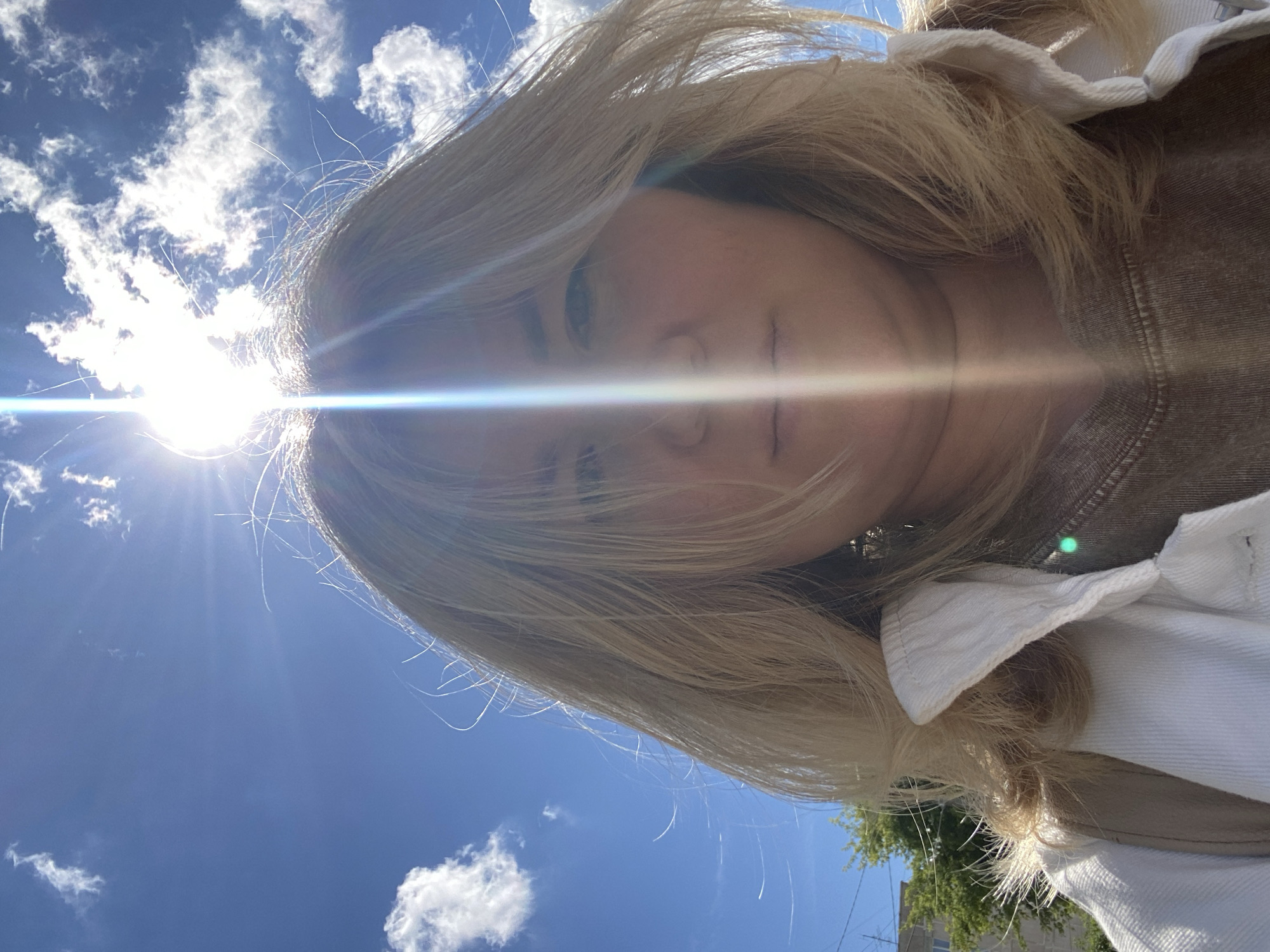 The image is a close-up, low-angle selfie of a woman with blonde hair taken outdoors against a bright blue sky with scattered white clouds. The sun is directly overhead and intensely bright, creating lens flare that runs vertically through the center of the image, obscuring part of the woman's face. She is wearing a white collared jacket over a brown garment. The background shows a building and some green trees, suggesting an urban or suburban environment. The context provided indicates the location is the city of Chaikovsky, Russia.
