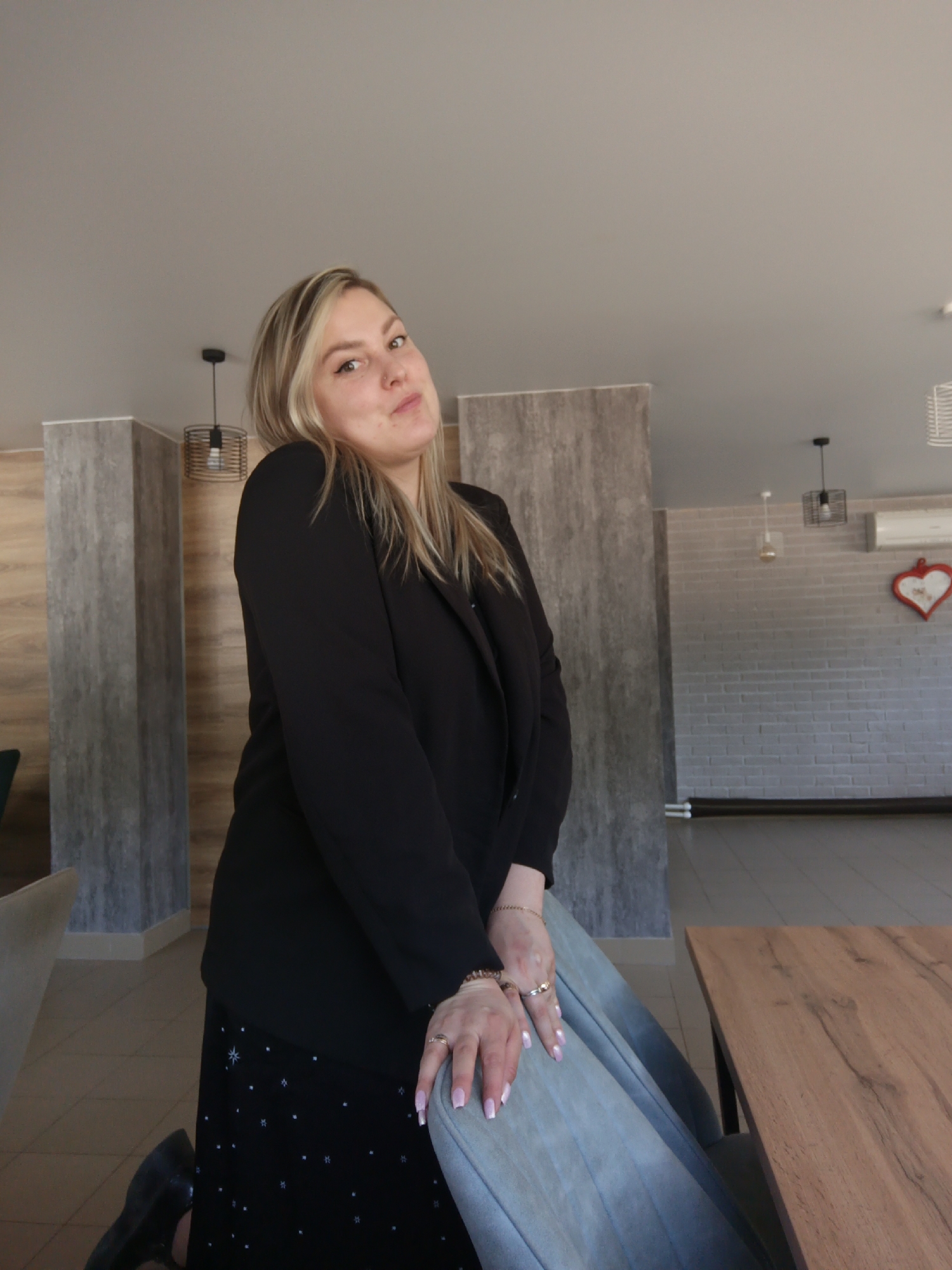 A woman with blonde hair is posing in a room. She is wearing a black blazer and a black skirt. Her hands are resting on a blue chair. A wooden table is next to the chair. The background features a white brick wall with a decorative heart, light fixtures, and a grey patterned wall. The location is Noviy, Russia.