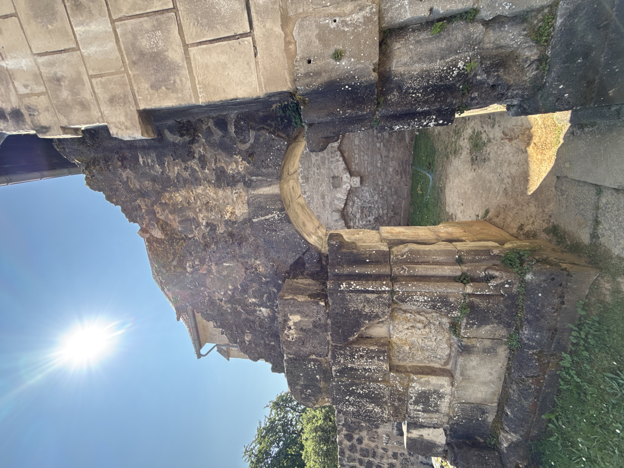 The image shows the exterior of the Romanesque former monastery church of Walkenried Abbey, located in Walkenried, Germany. The sun is bright and directly overhead, casting a strong glare in the upper left corner. The focus is on a weathered, arched stone entryway that leads into a courtyard. The stonework is ancient and shows signs of age and erosion, with moss and lichen growing on the surfaces. To the right of the arch, a wall made of lighter, cut stone blocks is visible. To the left, there is a stepped stone structure, also weathered and partially overgrown. In the background, through the arch, the courtyard appears to be a mix of dirt and grass. Trees and part of a building with a tiled roof are visible in the upper left quadrant. The scene suggests a historical site that is partially in ruins but maintained. There are no people visible in the image.