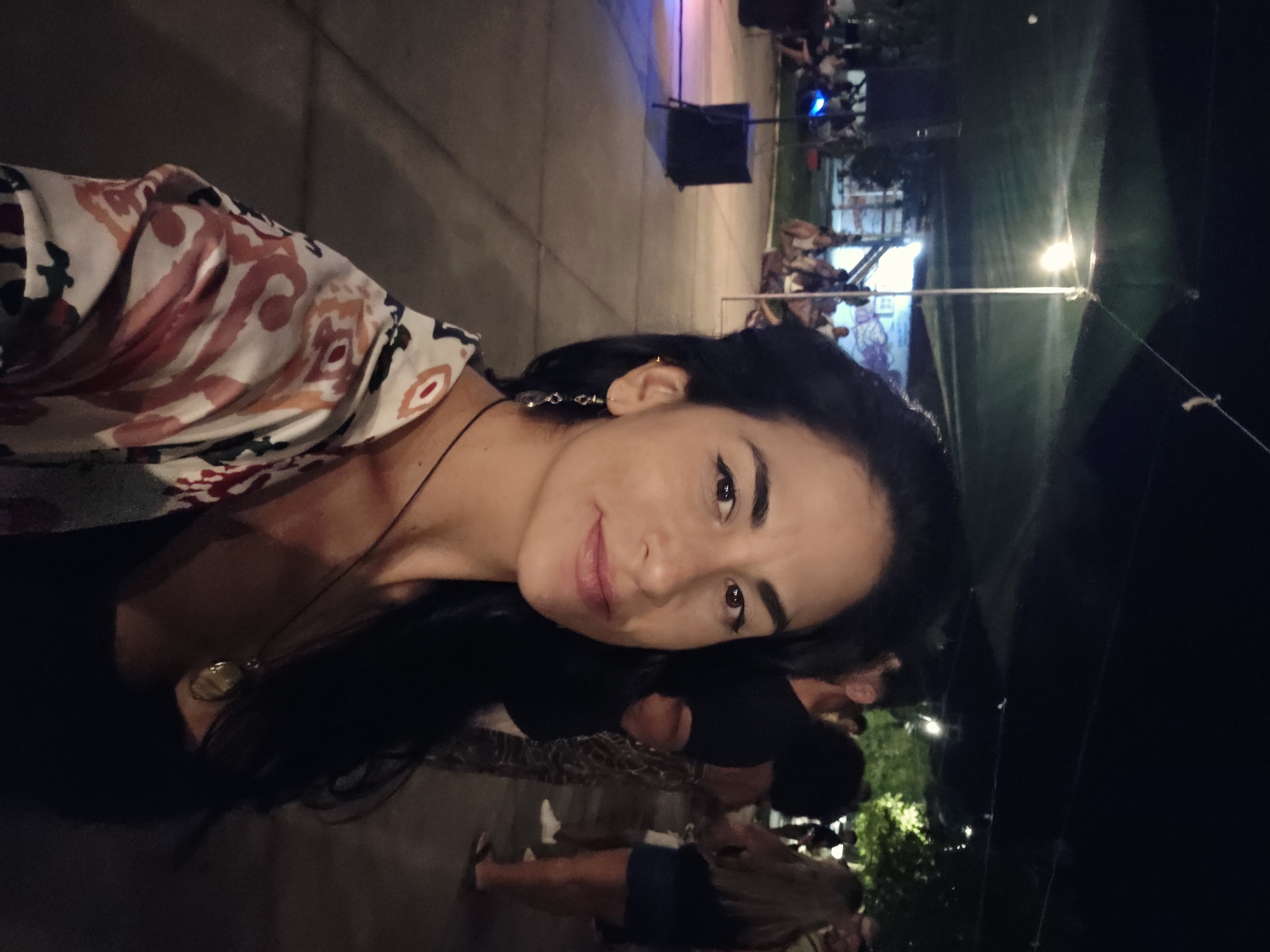 An outdoor scene at night depicts a medium-toned skinned woman, approximately 20-30 years old, with long dark hair, facing the camera for a close-up. She wears a dark top under an open white garment patterned with red, orange, and blue organic shapes. A thin dark necklace with an oval gold pendant is visible, along with a dangle earring and a small stud in her left ear. She exhibits a subtle smile.
The background reveals a social gathering under artificial illumination. An overhead dark canopy or tarp is visible, with string lights and a prominent incandescent bulb suspended by wires. The ground is paved. To the left, a dark speaker on a stand is illuminated by blue and purple light. Further back, numerous individuals are present; some are seated on the ground, while others stand and mingle. A light-skinned man with dark hair is visible directly behind the main subject's right shoulder, and a woman with long light-colored hair wearing a dark top and blue shorts is visible further right. Green foliage is scattered throughout the background, along with a building or wall displaying faint markings. The location is an outdoor public area within the city of Unknown, Unknown.