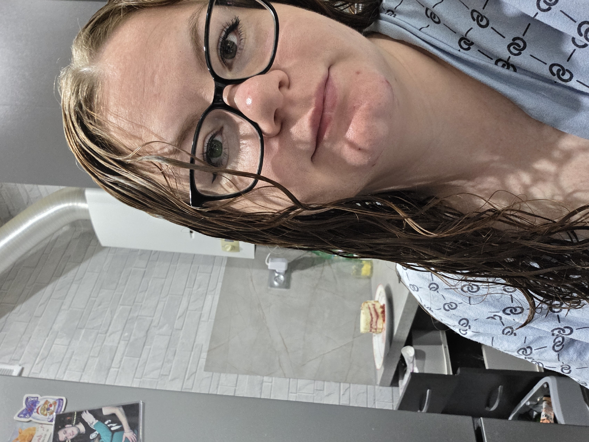 A close-up, slightly angled selfie shows a woman in a light blue robe with a repeating black pattern. She has wet, brown hair, dark-rimmed glasses, and fair skin with a few blemishes on her chin. In the background, on a kitchen counter, is a slice of layered cake on a white plate. The kitchen wall has white textured tiles, and a silver exhaust pipe is visible overhead. A photograph of a couple is attached to a gray refrigerator on the left side of the image. The scene appears to be indoors, likely in a residential kitchen. The city of Zheleznogorsk, Russia, is mentioned as the location, though no specific landmarks confirm this.