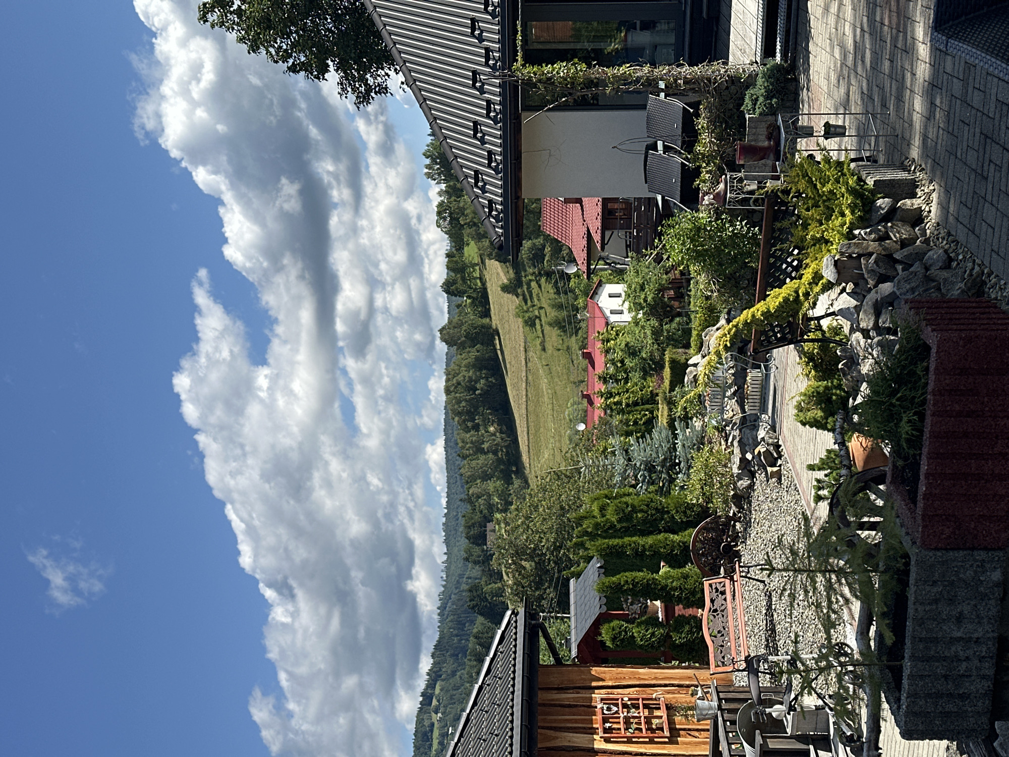 This image depicts a scenic view of a courtyard in Wisła, Poland. The foreground features a paved patio with stone planters and various decorative elements like benches, rocks, and small trees. The middle ground showcases a lush garden with numerous evergreen trees and bushes, leading to a cluster of houses with red-tiled roofs. In the background, rolling green hills covered in trees form a picturesque landscape under a bright blue sky dotted with white, fluffy clouds. The overall impression is one of a tranquil, rural setting.