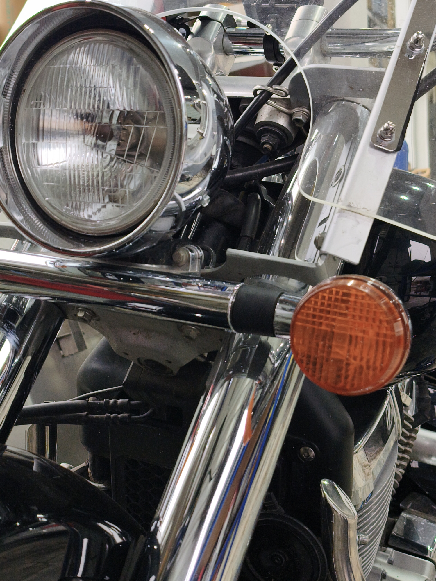 The image is a close-up, vertical shot of the front portion of a motorcycle, focusing on the headlight and a portion of the engine and handlebars. The motorcycle is predominantly silver and black, with a large, chrome-encased headlight dominating the upper left. To the right of the headlight, a clear windshield is visible, held by chrome supports. An orange turn signal is prominent in the lower right quadrant of the image. The motorcycle's engine components, exhaust pipes, and various chrome accents are visible throughout the frame. There are no people or discernible environmental details that would indicate a specific location within Noviy, Russia. The lighting is even, suggesting an indoor setting or an overcast day.