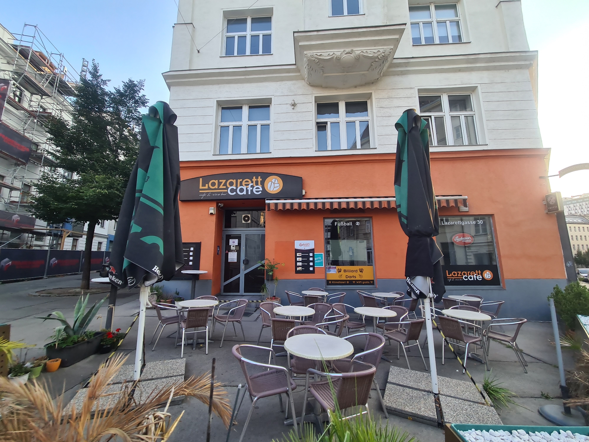 The image captures the exterior of a cafe named "Lazarett Cafe" located at Lazarettgasse 30 in Vienna, Austria. The cafe has an orange facade and an awning over the entrance. Outside the cafe, there are several tables and chairs set up, suggesting an outdoor seating area. Two large, closed umbrellas are present, one of which is dark green with a logo. In the foreground, there are potted plants and dried foliage. The building has multiple windows on its upper floors. To the left of the cafe, scaffolding is visible, indicating construction or renovation work on an adjacent building. The sky is clear and blue. No people are visible in the image.