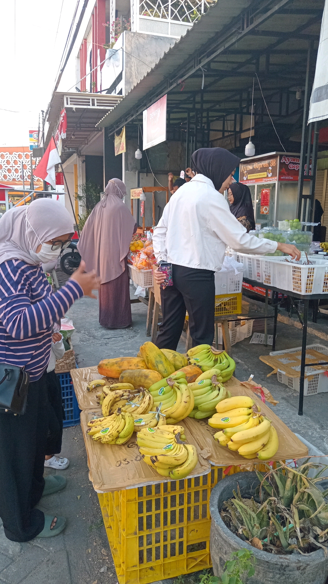 In Randuagung, Indonesia, a fruit stall is depicted. The scene is outdoors, with a paved surface beneath the stall. A variety of fruits, including bananas and papayas, are arranged on a table covered with brown paper, which rests on a yellow plastic crate. The fruits are displayed in bunches and individually. In the foreground, a person wearing a striped long-sleeved shirt, black pants, a face mask, and glasses is interacting with the stall, possibly making a purchase or inquiring about the fruit. The person is also carrying a black bag.
Behind the fruit stall, other individuals are visible. One person, wearing a dark headscarf and a white jacket, is reaching towards a white basket filled with fruit, possibly apples, on a table. Another person in the background is wearing a light brown headscarf and a long, dark dress. Further in the background, there are awnings and structures suggesting a market or street-side vendor area. A red and white flag, likely the Indonesian flag, is visible on a pole to the left. The overall impression is of a bustling local market.