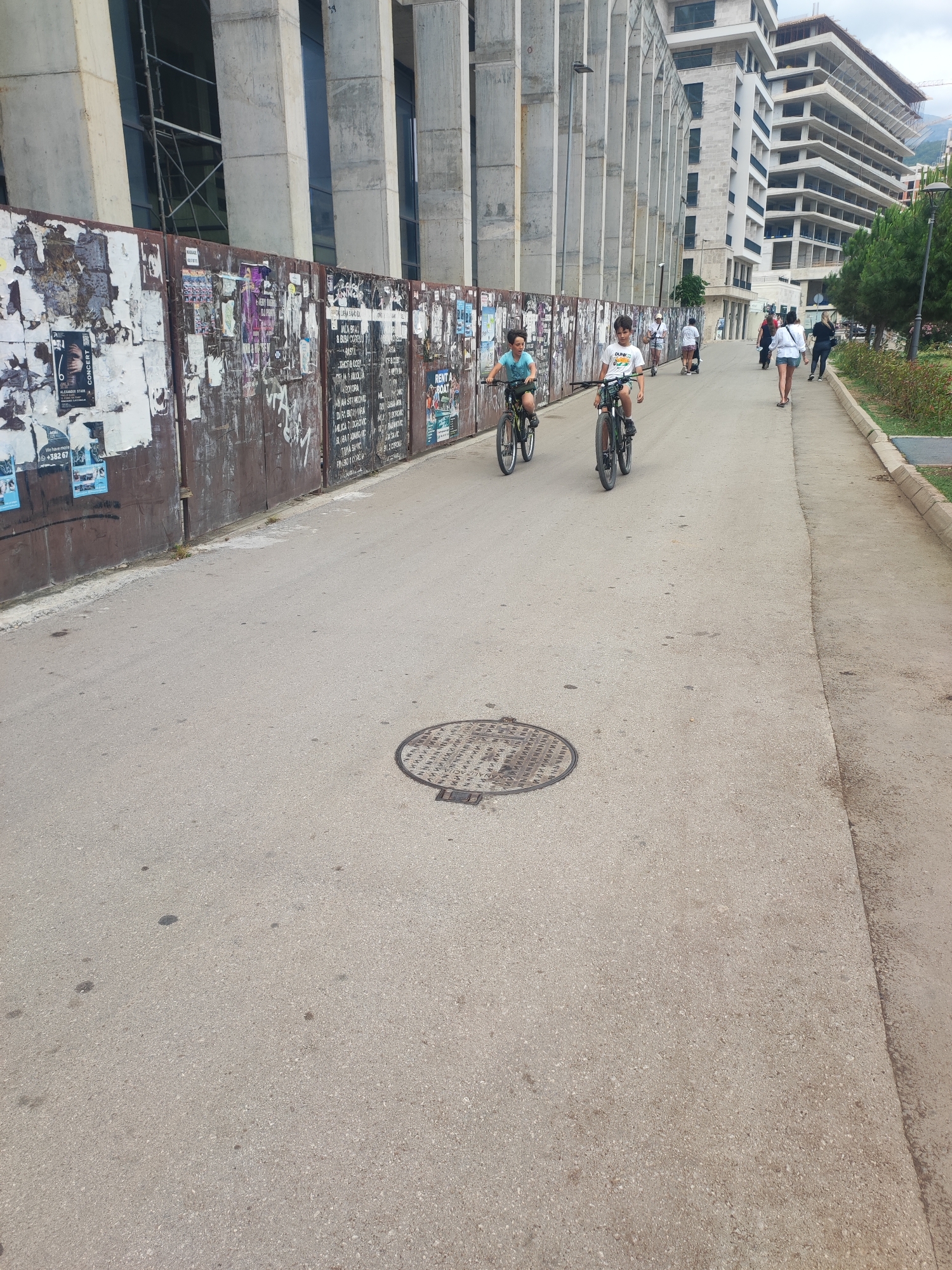An outdoor, eye-level shot captures a lively urban pathway, bathed in bright daylight under clear skies. Two boys, presumably pre-teens, are central to the image, riding bicycles on the asphalt path. The boy on the left wears a light blue t-shirt and dark shorts, while the boy on the right wears a white t-shirt with a graphic print and dark shorts.
To the left, a long, rusty metal construction fence stretches into the distance, heavily covered with numerous torn posters, flyers, and graffiti. Notable text includes "ALEXANDER STARK CONCERT" with a phone number "+382 67" and another sign saying "RENT A BOAT". Various other writings, some in Cyrillic script, are visible on the fence. Behind the fence, a large, modern building with tall concrete pillars and dark glass windows is visible, suggesting ongoing construction or recent completion. Further down the street, another multi-story concrete building appears to be under active construction.
To the right of the path, a narrower pedestrian sidewalk runs alongside a strip of green space with trees and shrubs. Several adults are seen walking along this sidewalk, some in the distance, one person appearing to be on an electric scooter or hoverboard. A manhole cover with a circular pattern is present on the asphalt in the mid-ground. The scene portrays typical daily activity in a developing urban area, likely in Budva, Montenegro, given the contemporary architecture and construction, common in growing coastal cities.