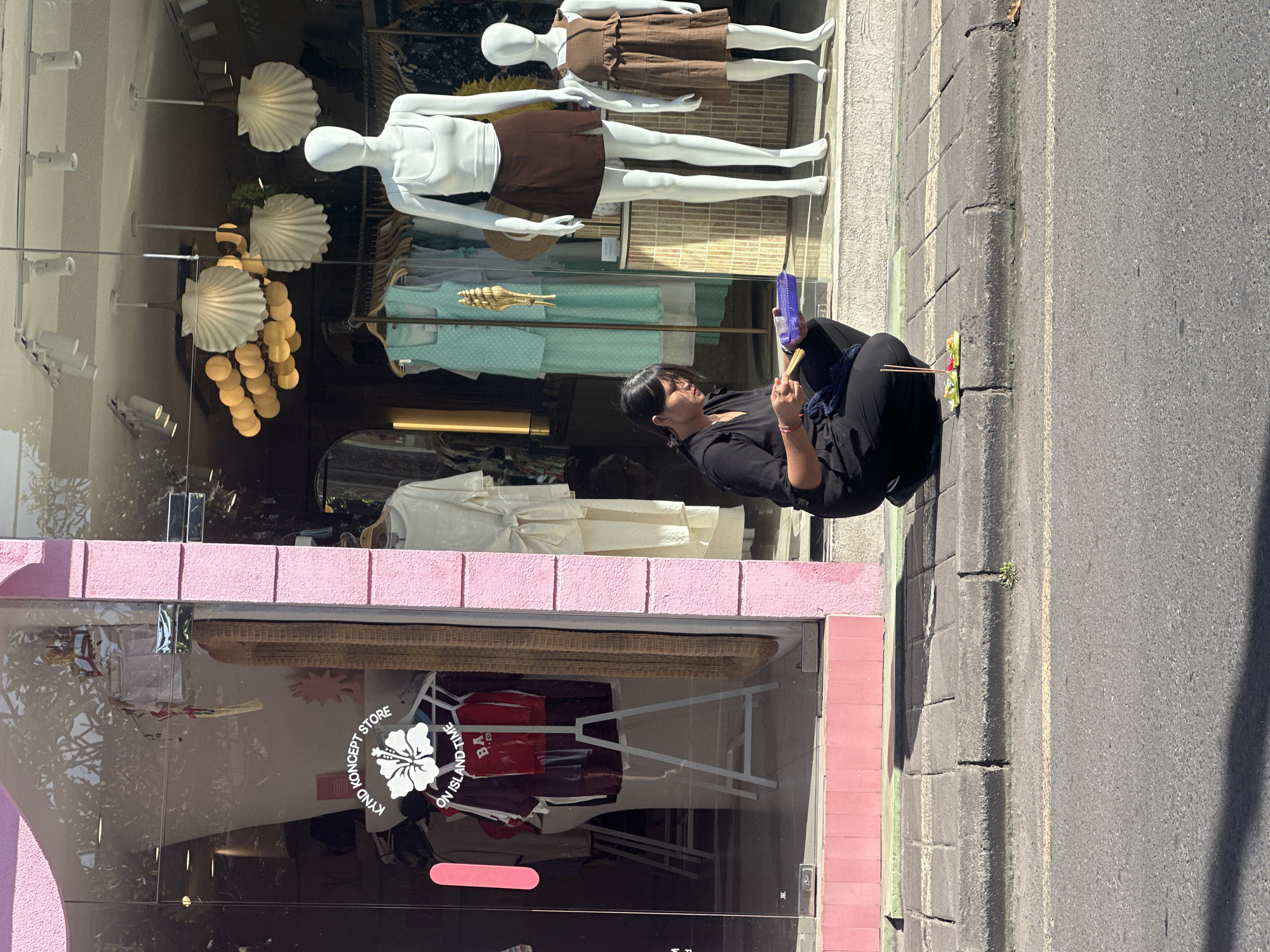 A woman is crouched on a sidewalk in front of a store in Ubud District, Indonesia, holding a purple tray and incense sticks. She is placing the incense sticks onto a small offering placed on the sidewalk. The store, painted pink, has a large glass window displaying mannequins and clothing. The sidewalk is made of concrete pavers and is bordered by a curb and a street. Sunlight casts shadows across the scene.