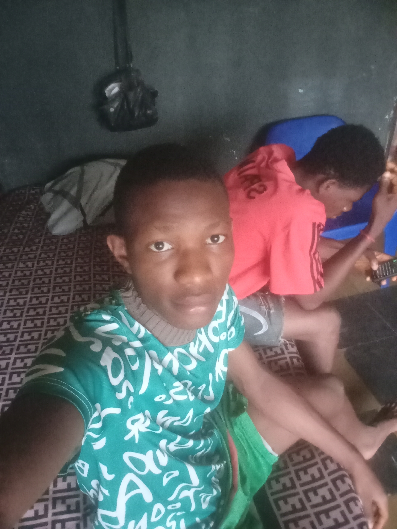 In an indoor scene, two young individuals are present. The individual in the foreground, facing the camera, is wearing a teal t-shirt with white lettering and green shorts. Their skin tone is dark, and they have short hair. The second individual, positioned in the background to the right, is wearing a red t-shirt and appears to be holding a cell phone in their right hand. They are seated on a blue surface. The environment features a patterned dark surface beneath the individuals, possibly a bed or couch, and a dark wall behind them with a black bag hanging from it.