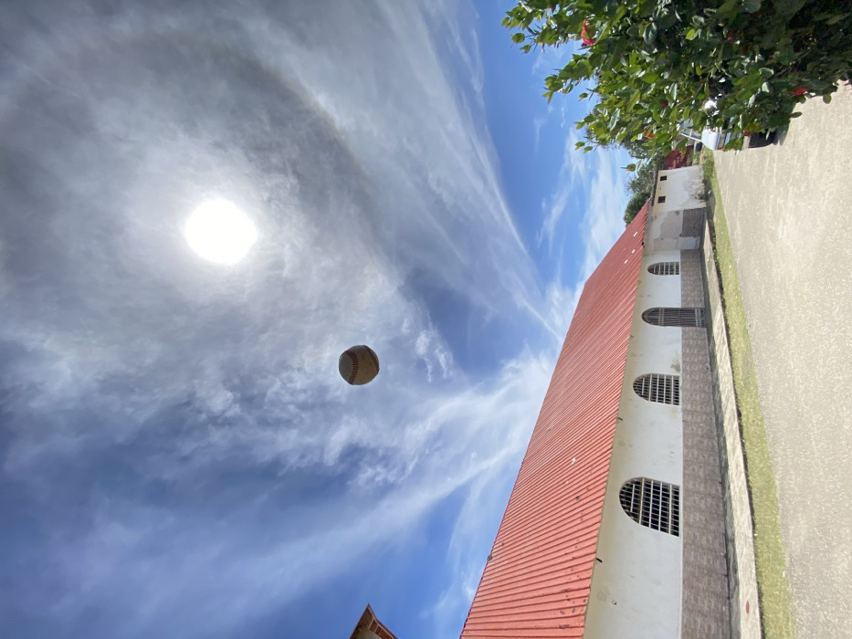 The image depicts a baseball suspended in mid-air against a bright blue sky with scattered cirrus clouds. A prominent solar halo encircles the sun, positioned in the upper right. Below the sky, a long, single-story building with a red corrugated metal roof and white walls occupies the lower left and central portion of the frame. The building features several arched window openings secured with white grates, and one arched entrance with a dark metal gate. A darker wainscot runs along the lower section of the building's facade. A narrow paved sidewalk runs alongside the building, followed by a strip of grass and a gray paved road extending into the foreground. On the right, a dense green bush with red accents partially obscures the front of a light-colored vehicle parked on the paved surface. The scene is set in Juan Griego, Venezuela.