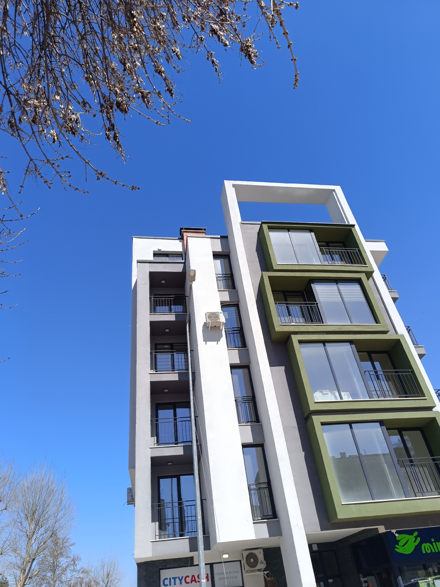 A low-angle, daylight shot shows a modern apartment building in Pleven, Bulgaria, characterized by white, gray, and olive green geometric shapes framing large windows and balconies. The building appears to have commercial spaces on the ground floor and residential units above. A storefront with a green sign is partially visible at the bottom right. Air conditioning units are attached to the exterior walls. Branches of a bare tree are visible in the top left and bottom left corners of the image against a clear blue sky. A sign with the word "CITYCASH" is visible at the building's base.