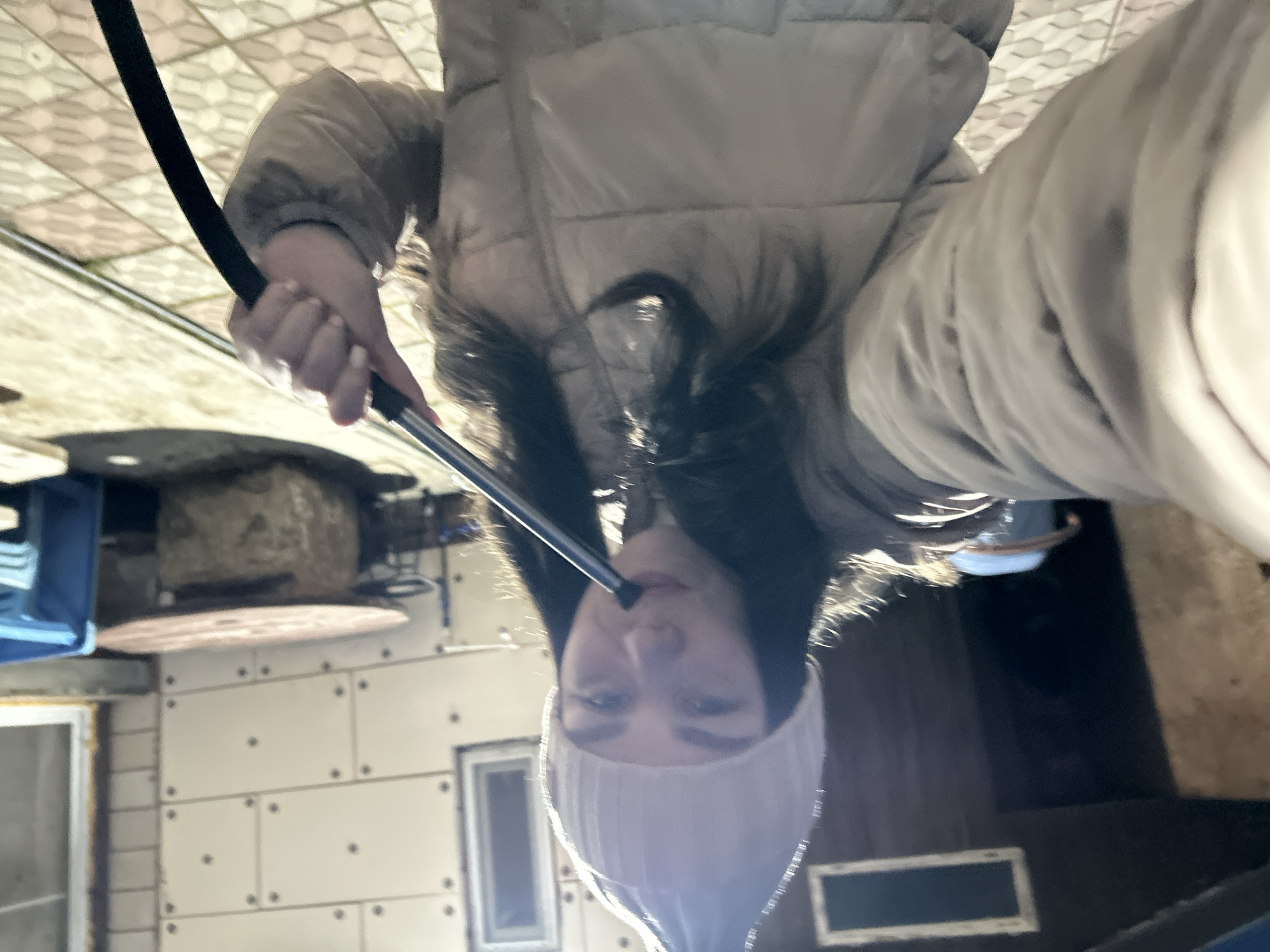 The image captures a young person, likely female, engaged in smoking a hookah. The photograph is taken from an inverted perspective, showing the subject and background upside down. She has long, dark hair and is dressed in a light-colored puffer jacket and a light-colored beanie. Her left hand holds a black hookah hose with a silver mouthpiece, which she is bringing to her lips.
The setting appears to be an informal indoor or covered outdoor space. In the background, from the inverted view, a geometrically patterned tiled surface resembles a ceiling. Below this, there are light-colored wall panels with visible fasteners and a rustic stone or brick structure, possibly a fireplace or oven. A blue object is partially visible on the far left. The lighting suggests it is daytime. The person's expression is neutral and focused on the activity. No visible text is present in the image. The scene suggests a casual and relaxed atmosphere. The location context is Фоки, Russia.