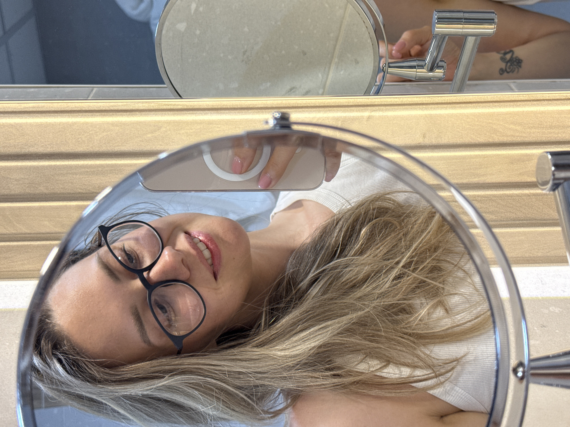A woman with blonde hair and glasses is taking a selfie in a bathroom mirror. She is wearing a white tank top. Her phone is visible in the mirror, and her hand with pink nail polish is holding it. There is a tattoo on her arm, visible in the reflection of another mirror. The bathroom appears to be modern, with a vertical wood-paneled wall and metallic fixtures. The location is identified as Nebug, Russia.