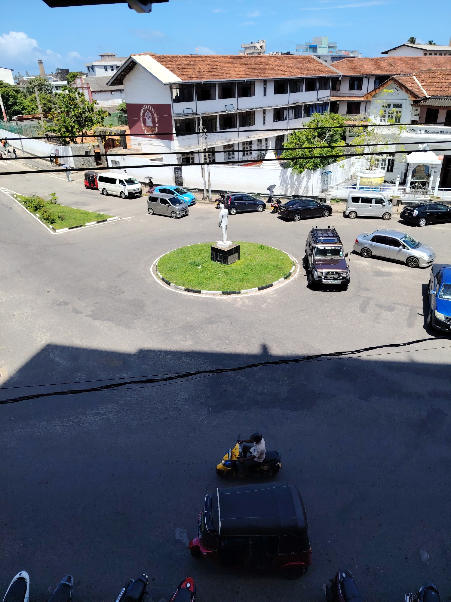 The image captures a street scene in an urban environment under a clear blue sky. The foreground shows a paved road with a shadow cast from a building. In the middle ground, several vehicles are present, including a yellow scooter with a rider, a black three-wheeled auto-rickshaw, and a dark-colored vehicle. Further back, a traffic circle is visible with a statue in its center. Around the traffic circle, more vehicles are parked and moving, including vans, sedans, and a rugged SUV. Buildings line the street, with one building in particular featuring a prominent red-tiled roof and a large emblem on its facade. The overall scene depicts a busy intersection or public space in a city, with elements of transportation and architecture.