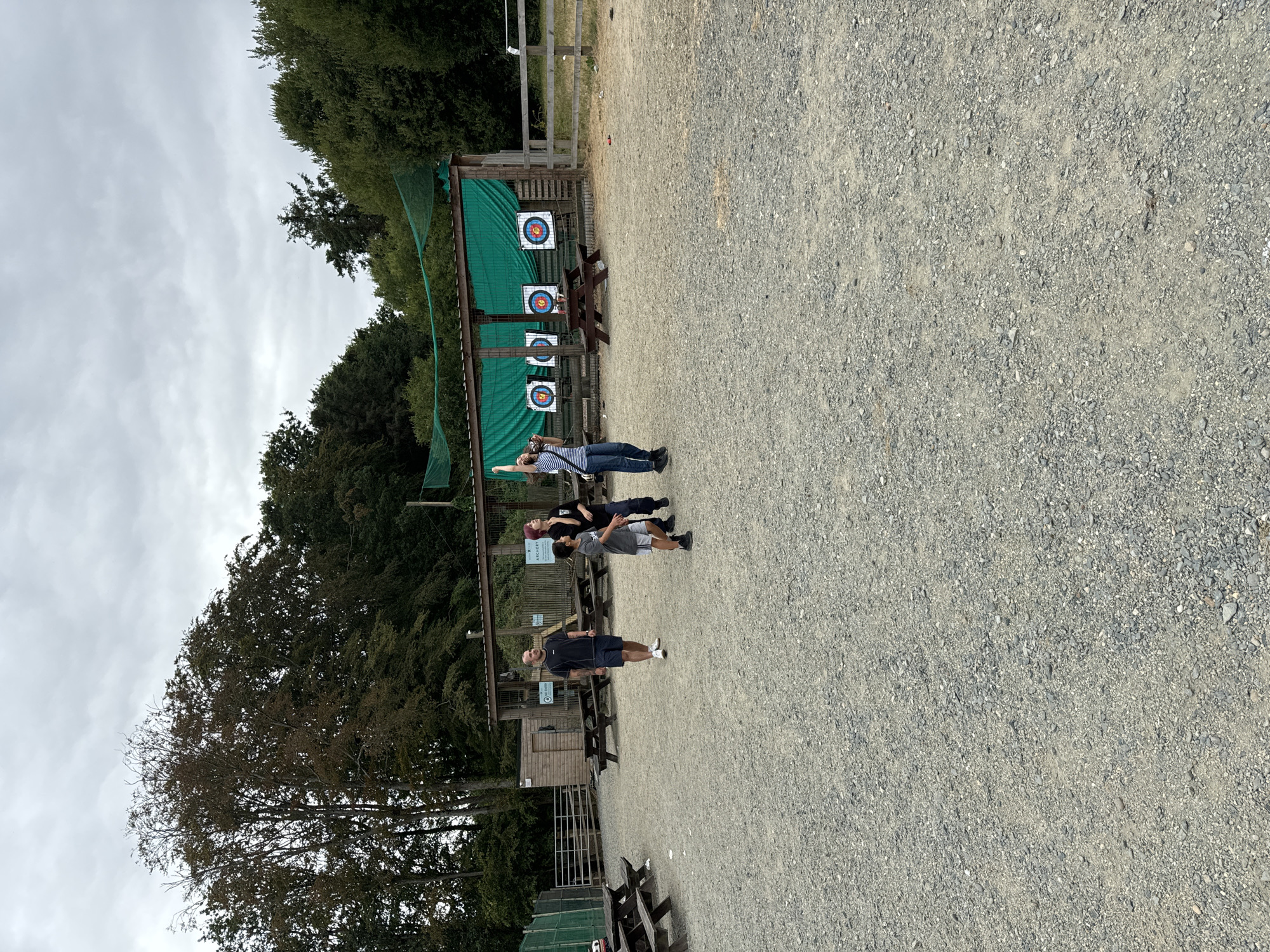 This image depicts an outdoor archery range, likely part of an adventure park or activity center, situated in the vicinity of Courtown, Ireland. The ground is covered with gravel. In the background, there is a wooden structure with a green netting overhead and several archery targets mounted on it. Three individuals are present. On the left, a man wearing a dark t-shirt and shorts stands looking upwards. To his right, a boy in a grey t-shirt and shorts is also looking up. Further to the right, a woman in a striped t-shirt and jeans is captured mid-jump, with her right arm extended upwards, suggesting she might be participating in or observing an activity. The surrounding area is bordered by trees and fencing.